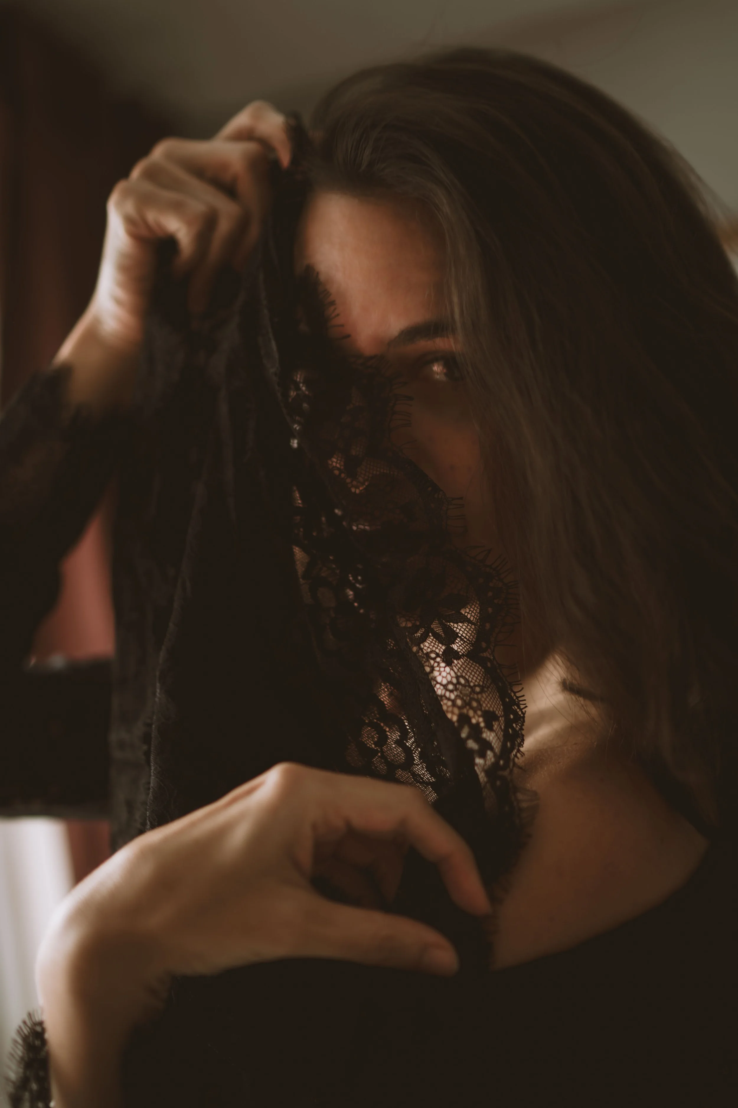 A woman partially hides her face behind black lace fabric, with one eye and part of her face visible, dark hair, and a hand forming a heart shape on her chest.