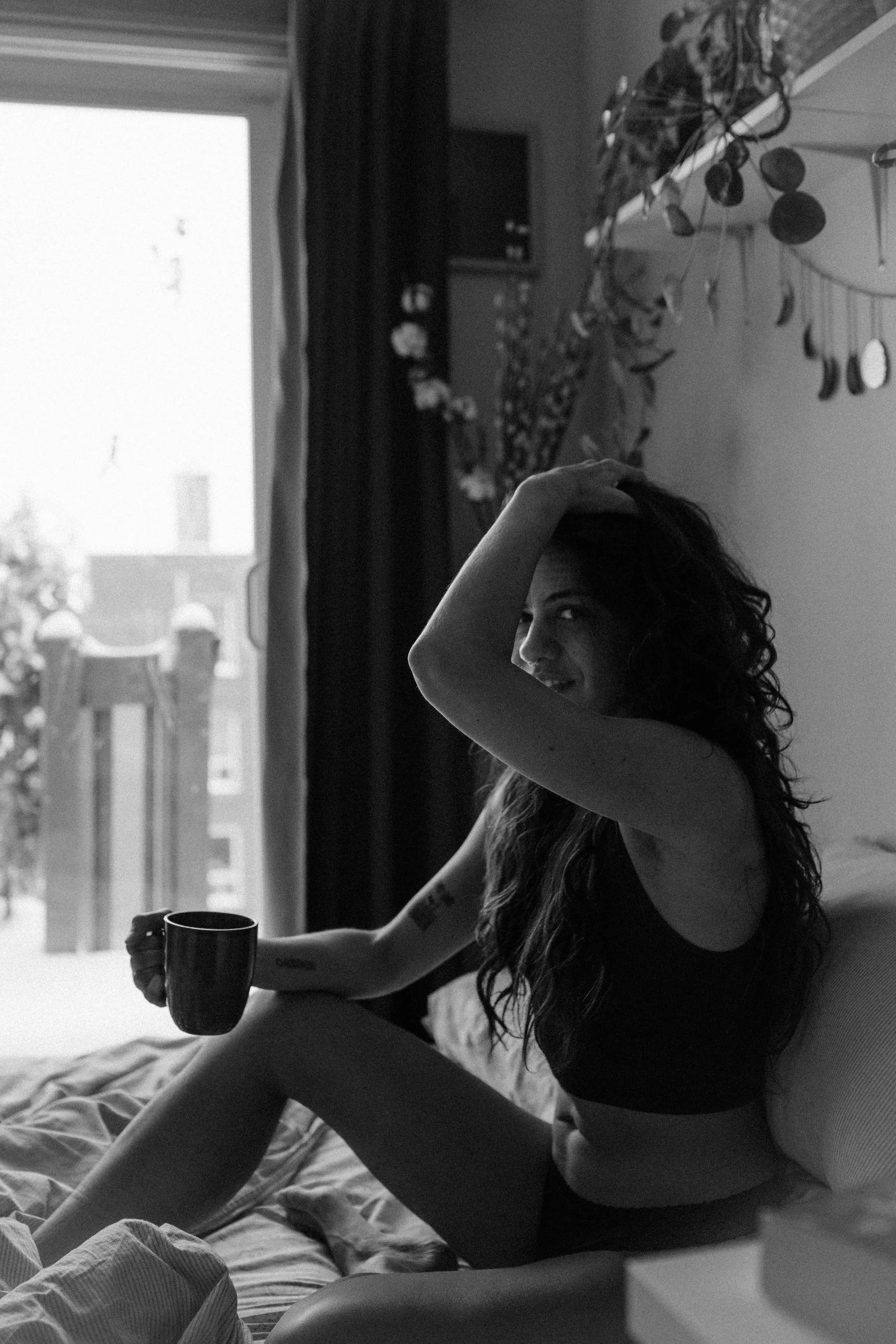 A woman sitting on a bed, holding a mug in her right hand, with her left hand resting on her head, smiling at the camera, in a room with a window, curtains, and wall decorations.