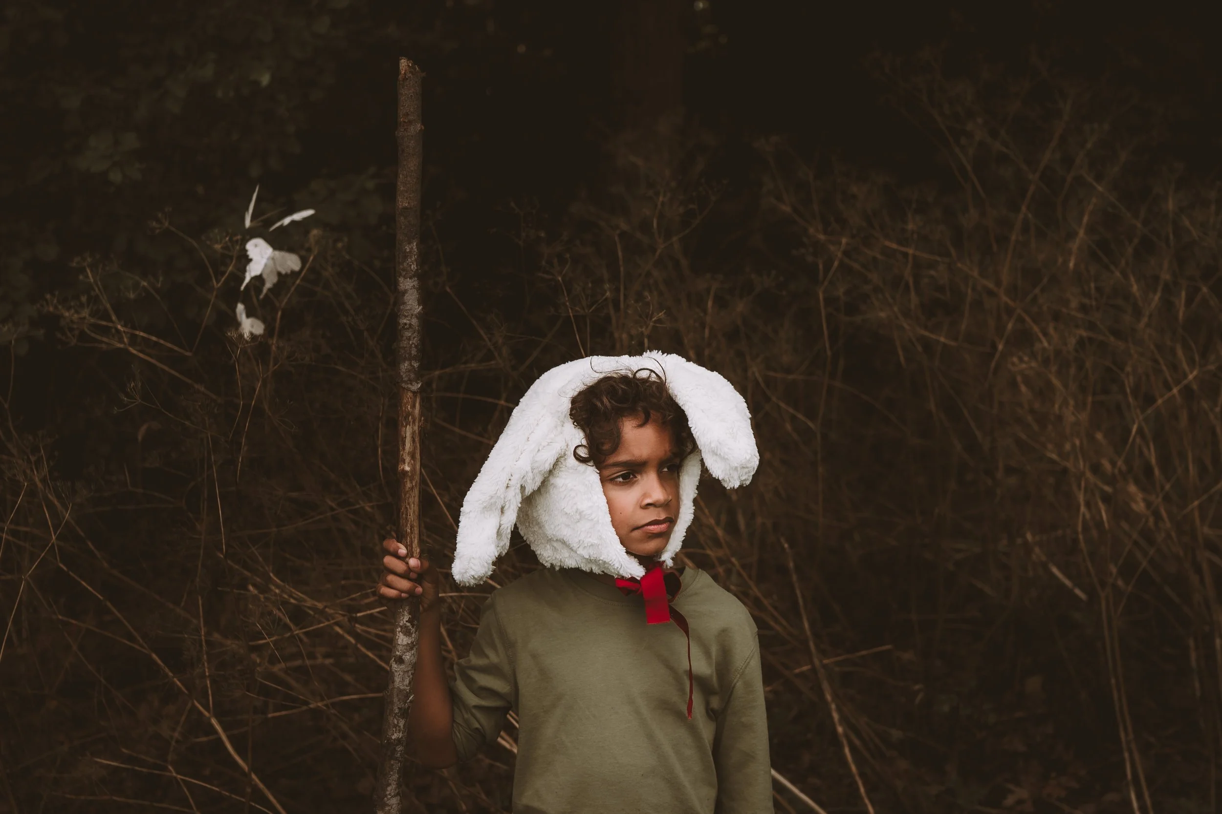 A young boy with curly hair wearing a white furry hood that resembles a bunny, holding a stick, standing outside in a natural setting with dry branches and dark foliage.