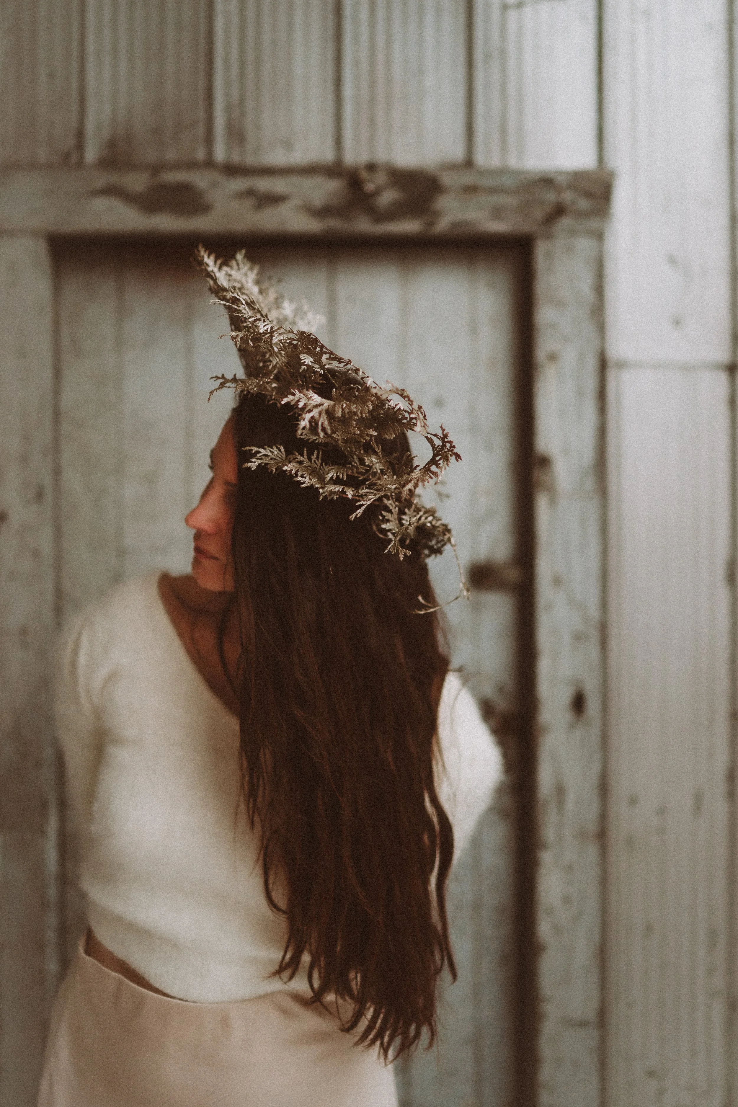 Woman with long dark hair wearing a white knit sweater and a crown made of dried plants, standing against a rustic wooden background.