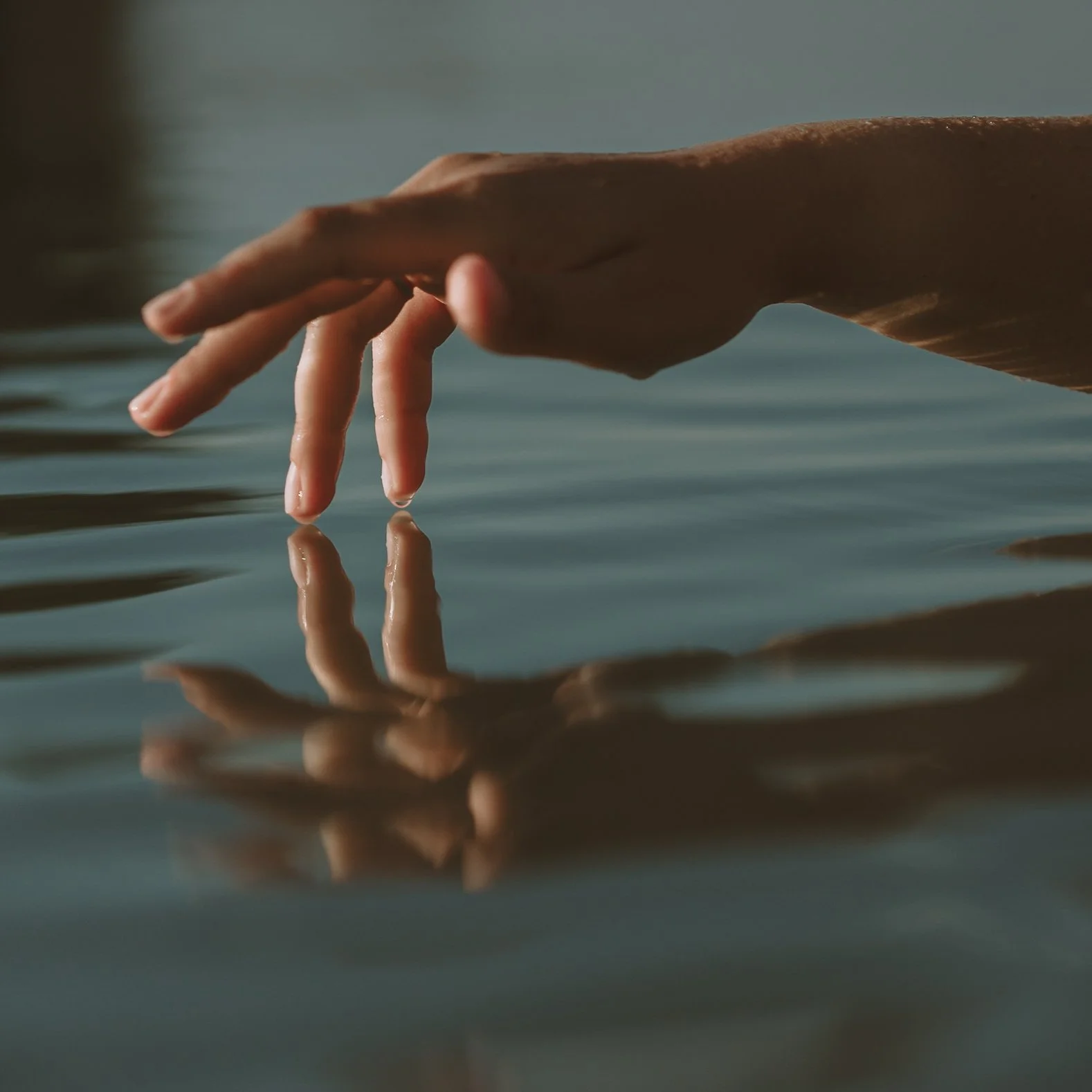 A close-up of a person's hand touching the surface of water, with water droplets on their fingers, and the reflection of the hand visible in the water.