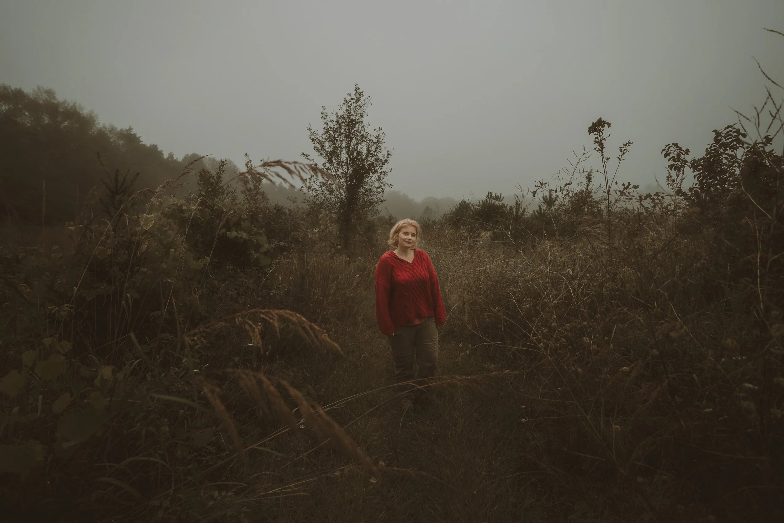 A woman with blonde hair wearing a red sweater and gray pants standing in a foggy, overgrown outdoor area with trees and bushes.