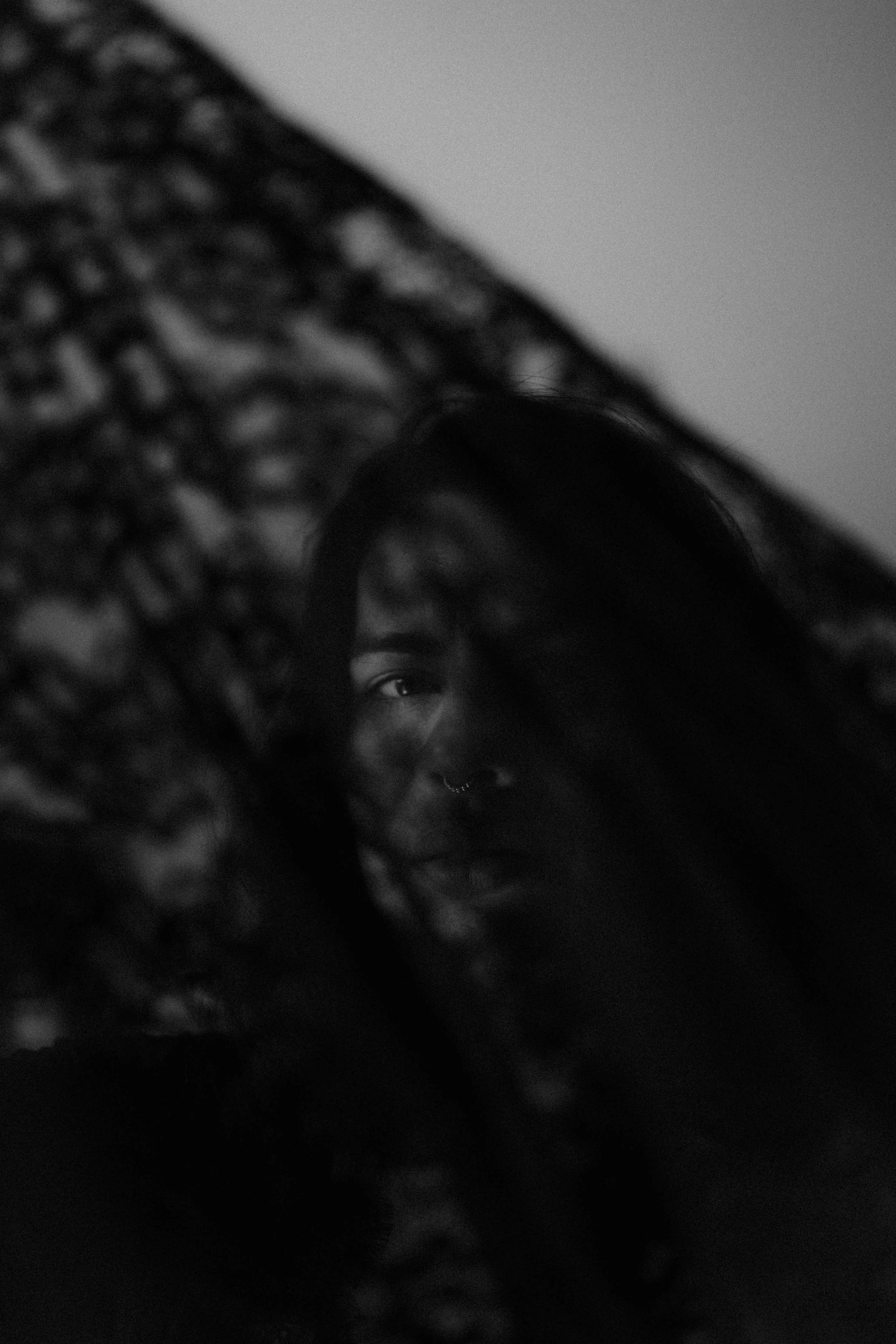 Black and white photograph of a young woman with dark hair and a nose ring, partially obscured by a patterned lace fabric.