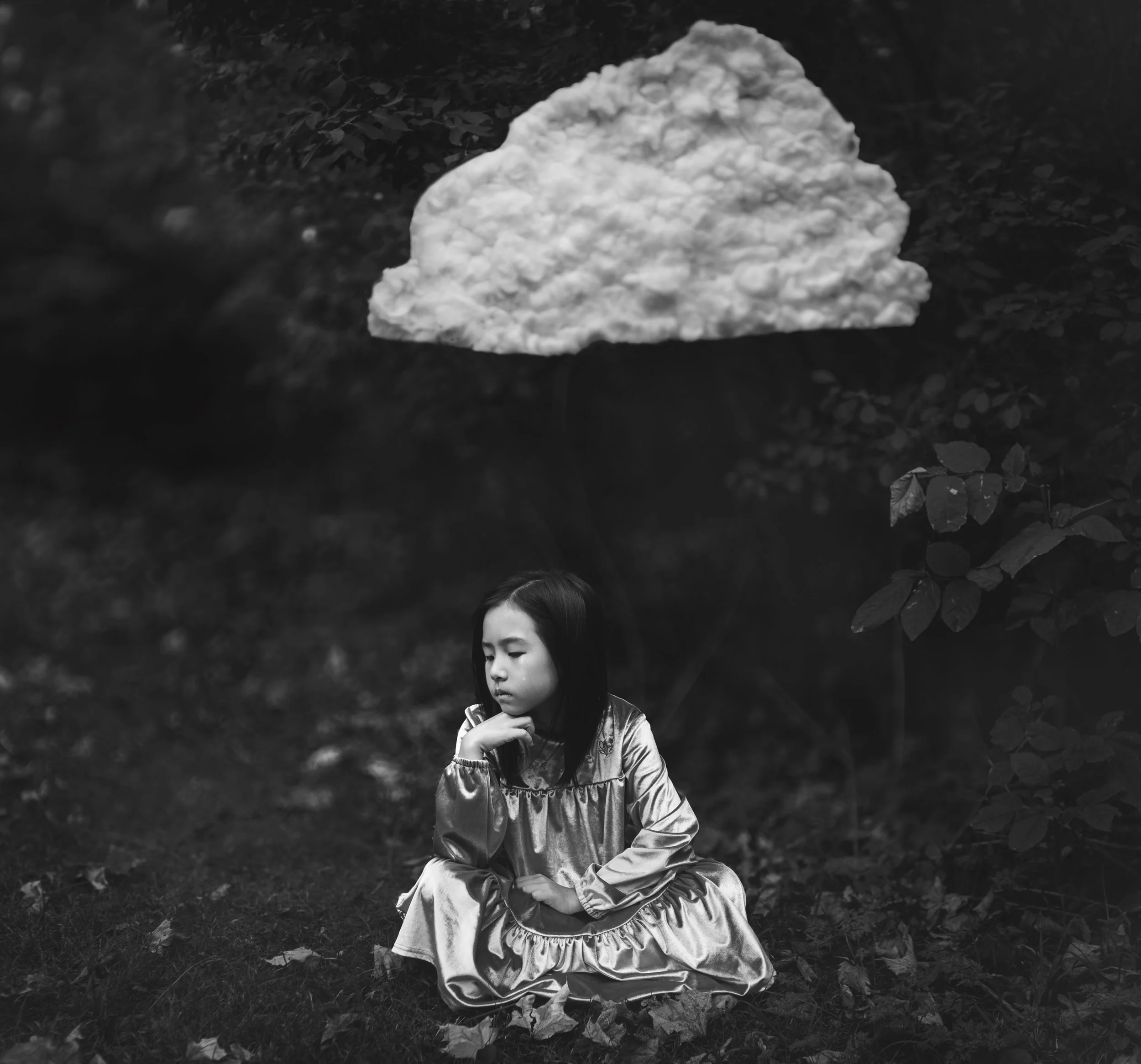 Black and white photo of a young girl sitting on the ground in a forest, wearing a satin dress, with a contemplative expression, and a large cloud-like shape floating in the sky above her.