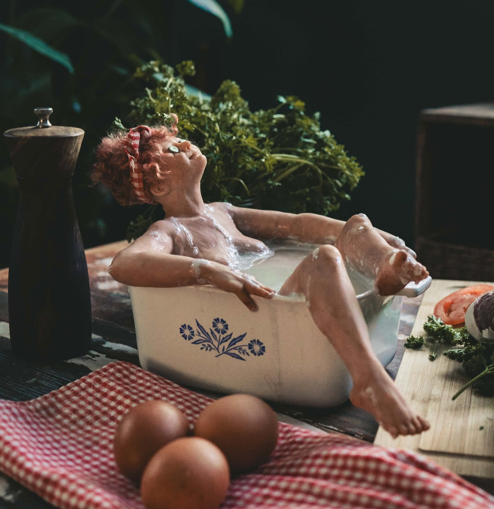A sculpture of a woman with red hair and a red bandana taking a bath in a kitchen dish with floral design, surrounded by kitchen items and vegetables, including eggs, a chopping board with sliced tomato, and fresh herbs.