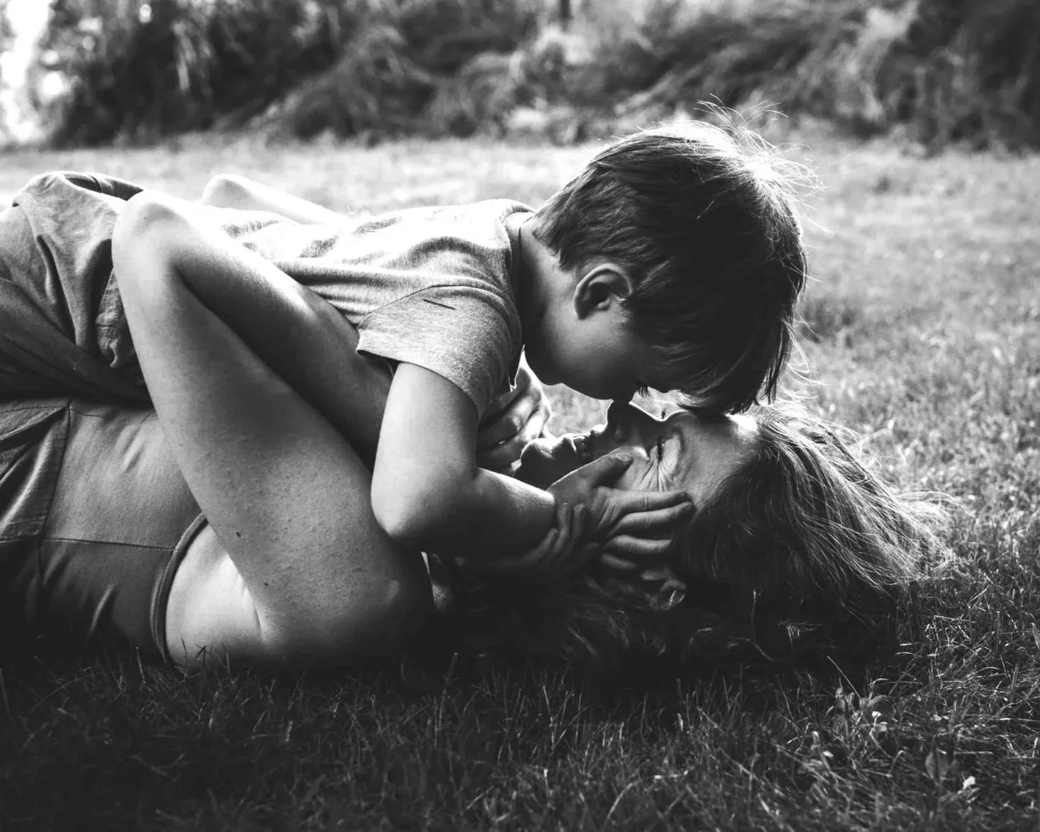A young boy and an adult woman are playing on the grass outdoors. The boy is lying on top of the woman, face close to hers, with his hands on her face. Both are smiling and appear to be enjoying a tender moment.