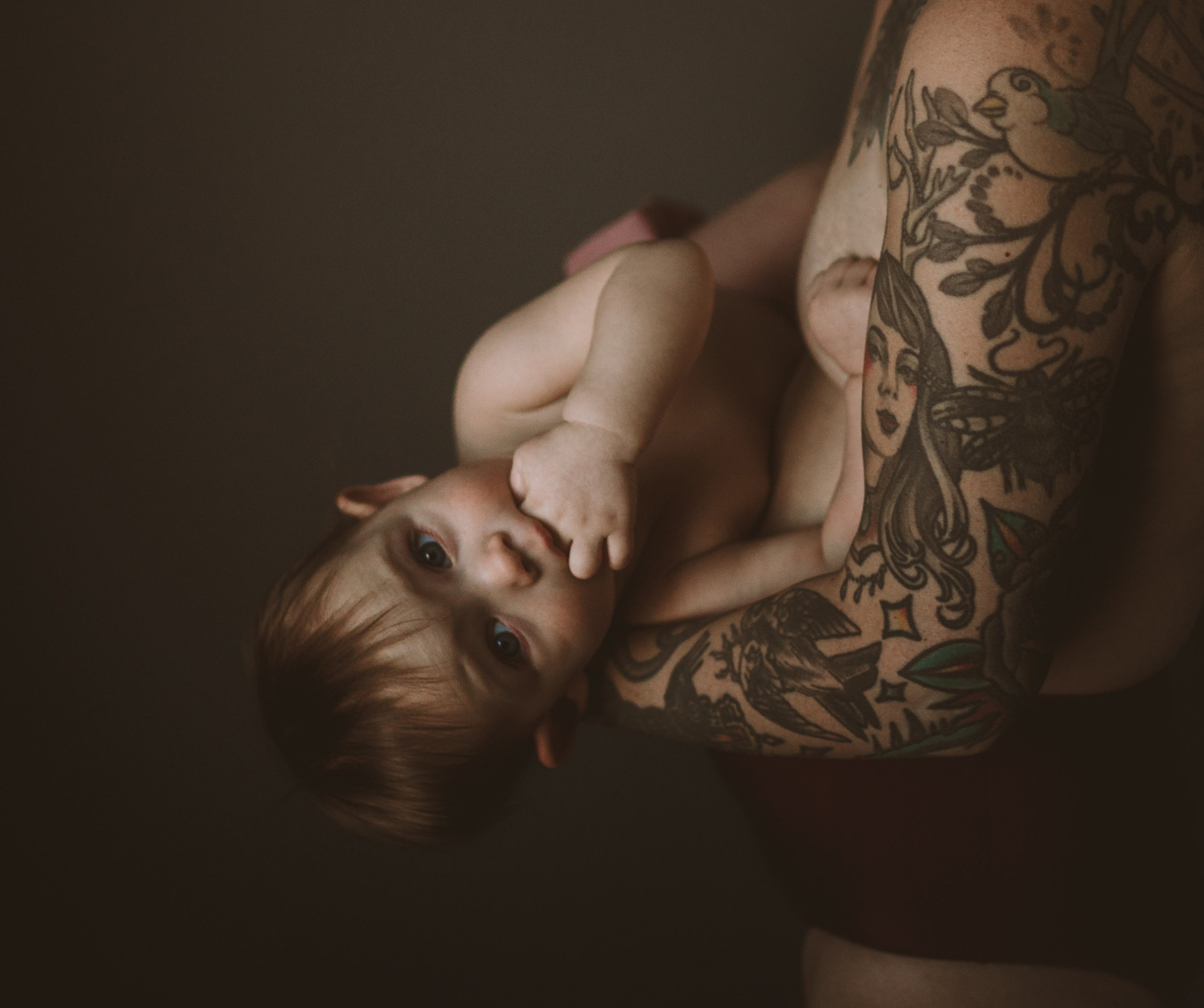 A person with extensive tattoos on their arm and hand is lying down, holding a small dog close to their face. The dog has a light-colored coat and is looking up at the person.