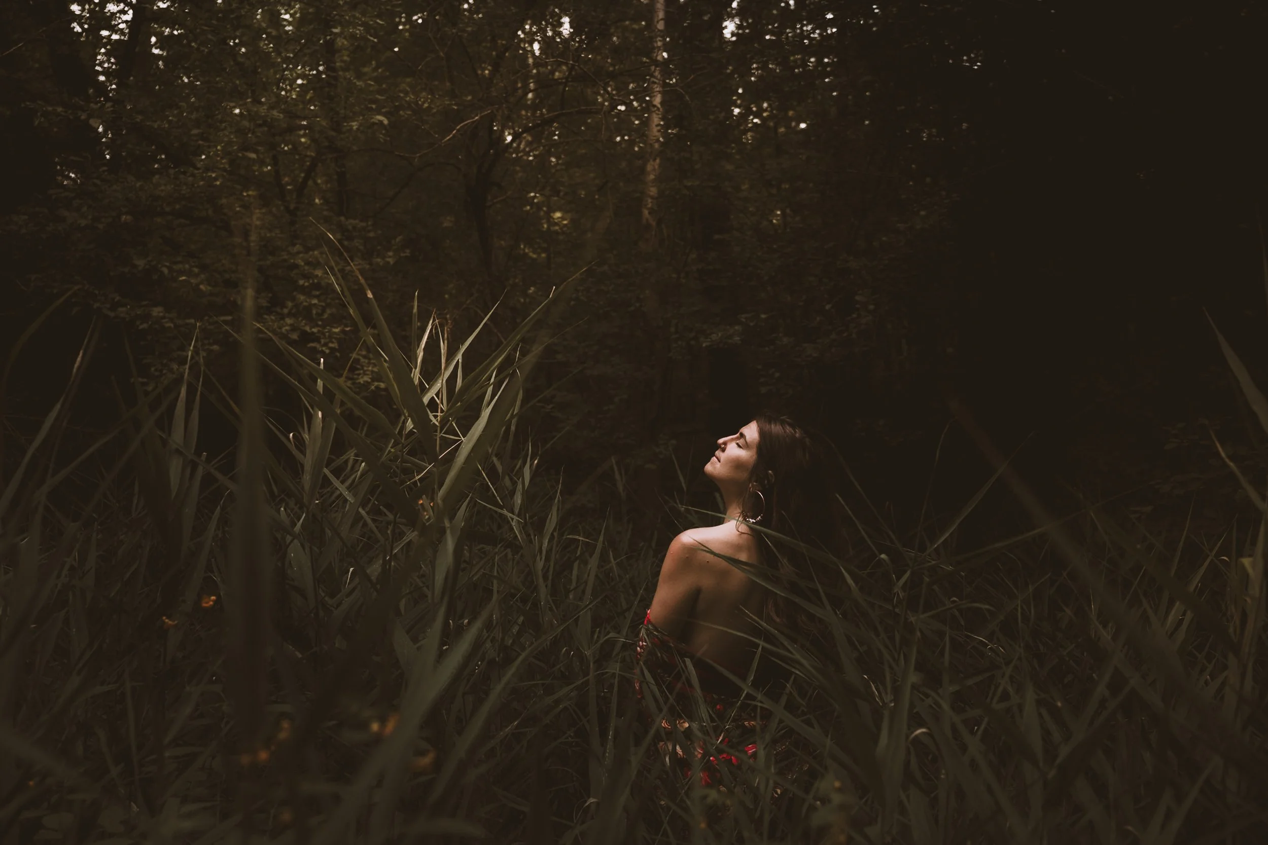 A woman with dark hair and large hoop earrings standing in tall grass at night, with a dark wooded background.