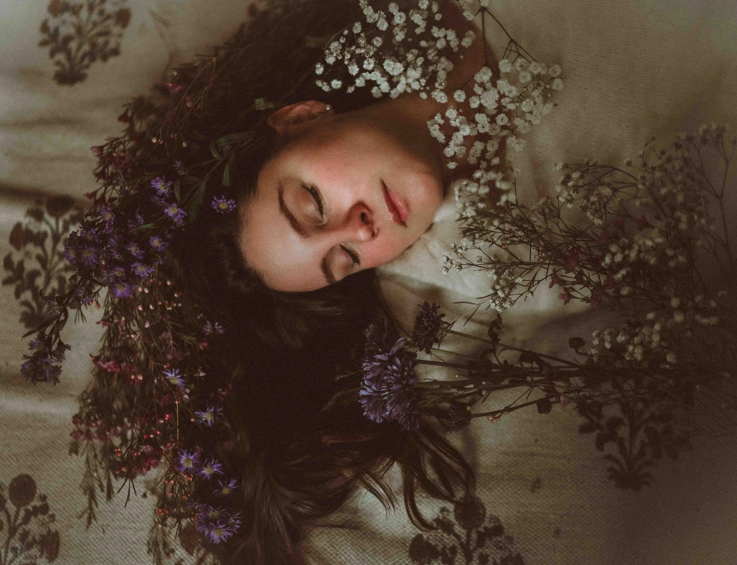 A woman with long dark hair and closed eyes lies on a beige fabric, surrounded by various white, purple, and pink flowers.