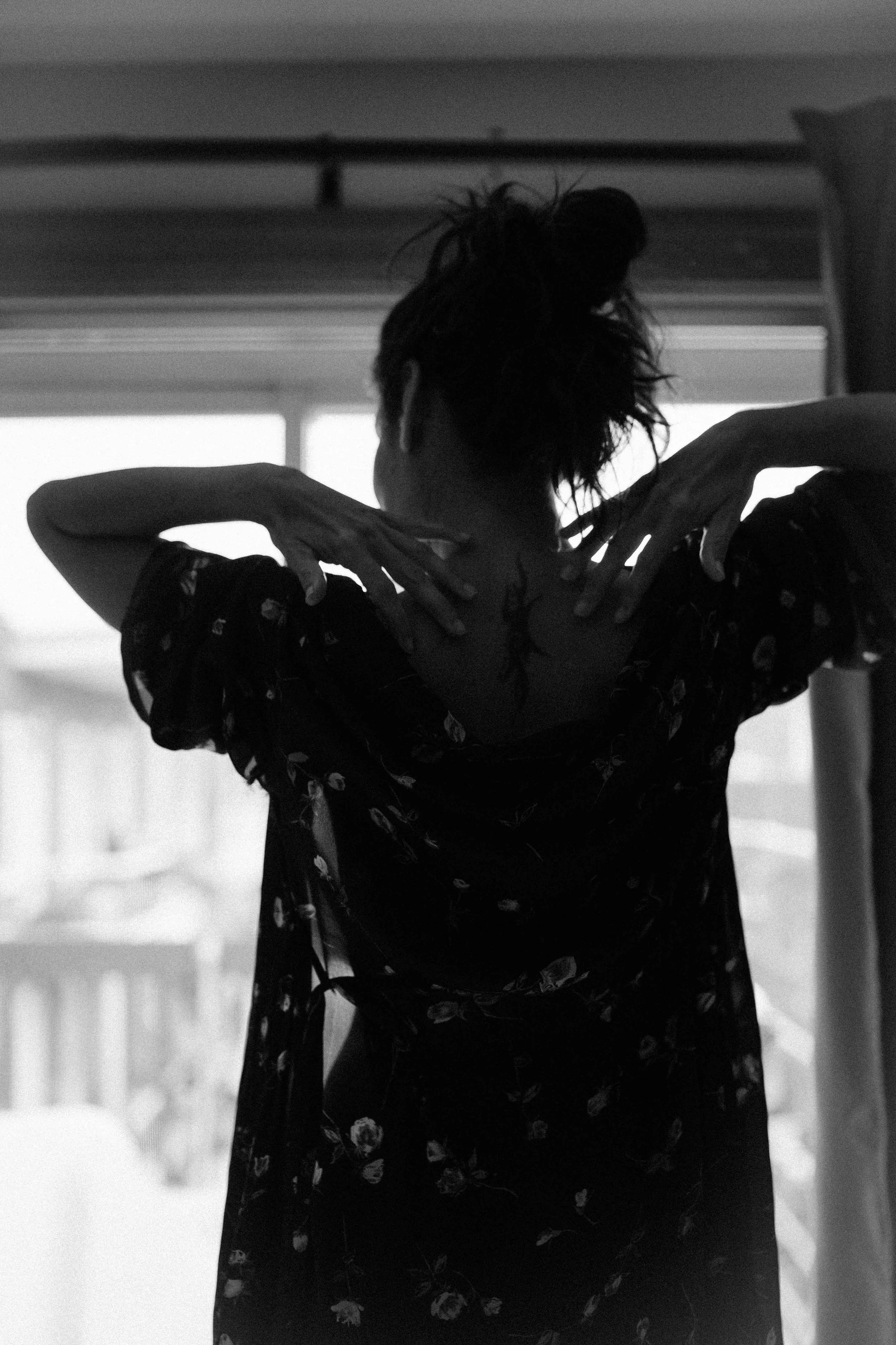 Black and white photo of a person with messy hair, wearing a floral dress, standing in front of a window with a landscape view, their back to the camera, hands raised near their neck.