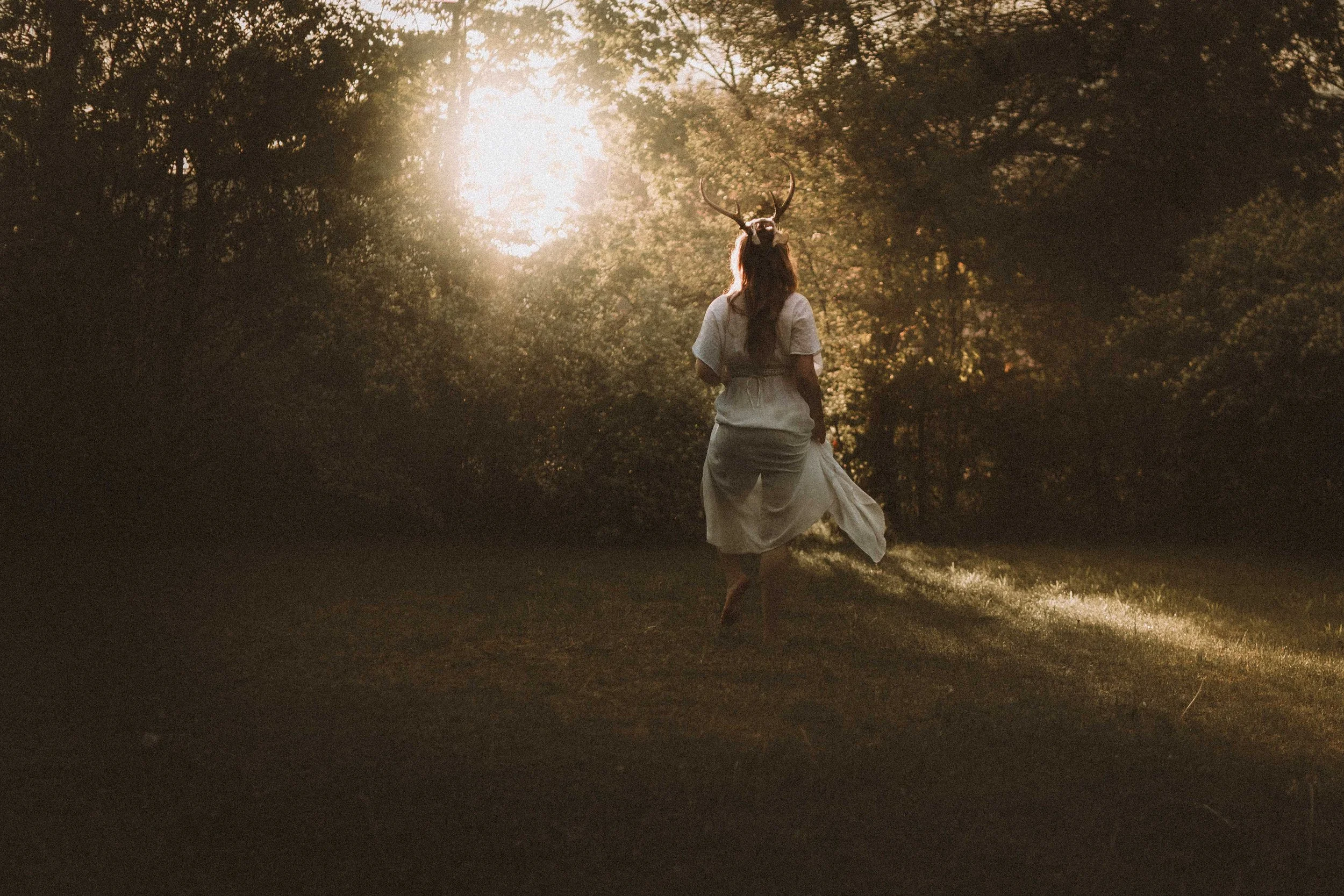 A woman dressed in a white dress, wearing a deer antler headpiece, walking through a forest at sunset with sunlight filtering through the trees.