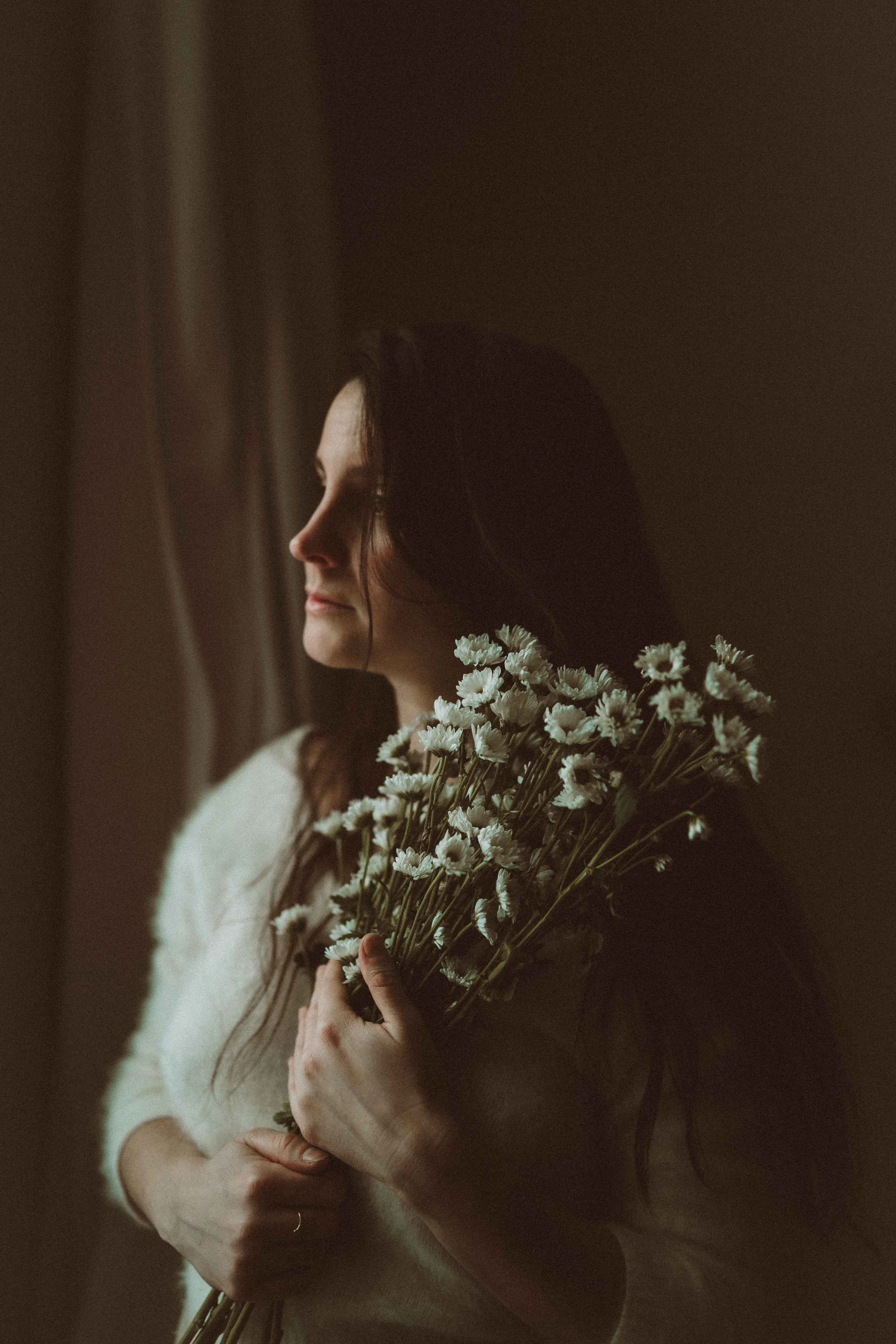 A woman with long dark hair holding a bouquet of white daisies, standing near a window with soft natural light.