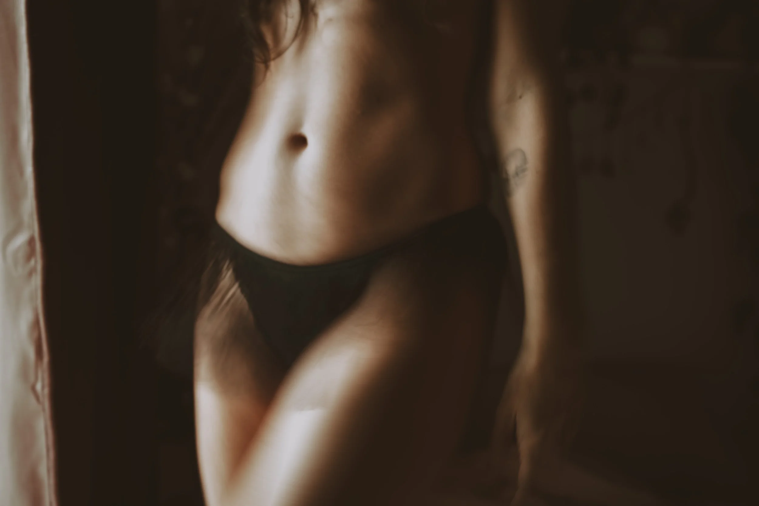 A close-up photo of a woman's midsection, showing her bare abdomen and black underwear, with a tattoo on her upper arm.