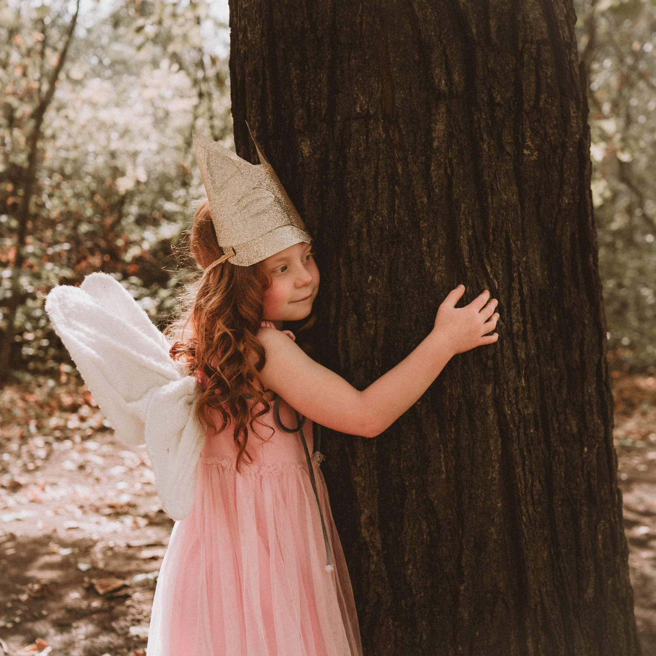 A young girl with long red hair, wearing a pink dress, hugging a large tree in a woodland area with fallen leaves on the ground.