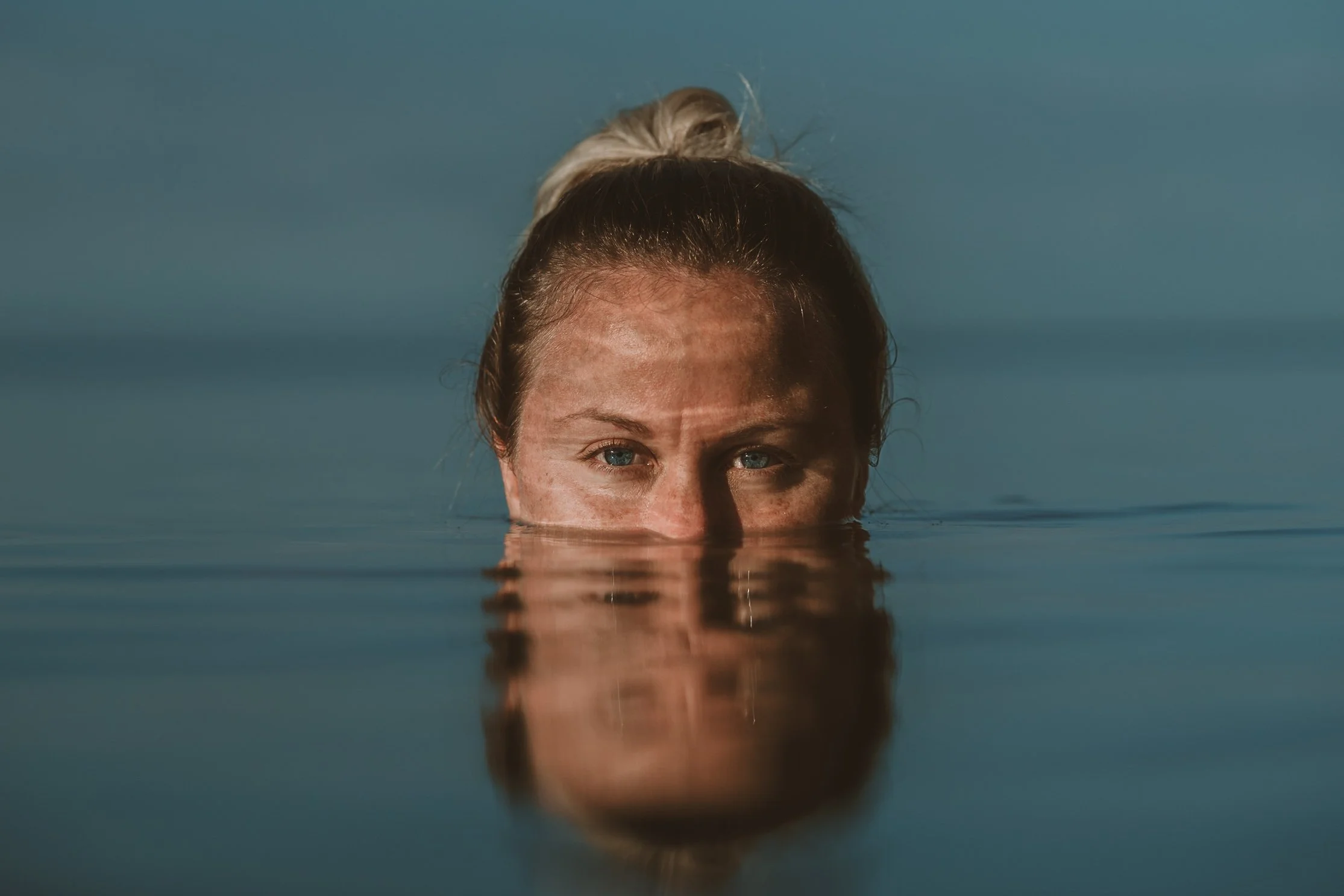 Close-up of a woman's face with blue eyes, partially submerged underwater, with her hair tied in a bun. The water covers her nose and mouth, reflecting her eyes and forehead.