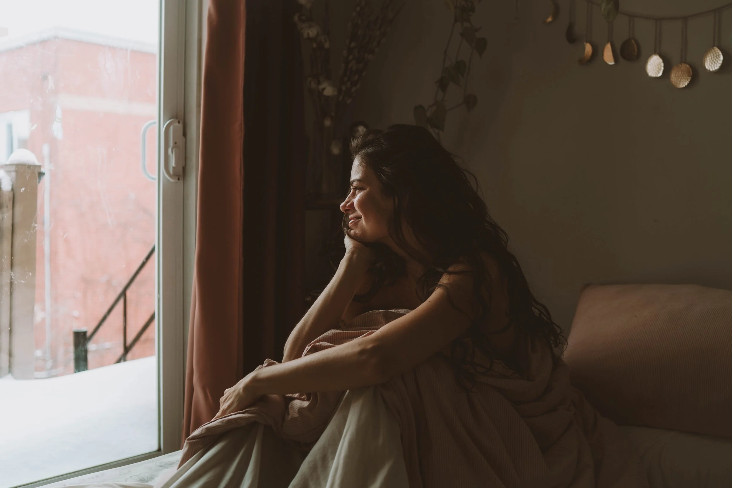 A woman with long dark hair sitting on a bed near a window, looking outside with a smile. The room has a wall decorated with hanging circular ornaments and floral artwork.