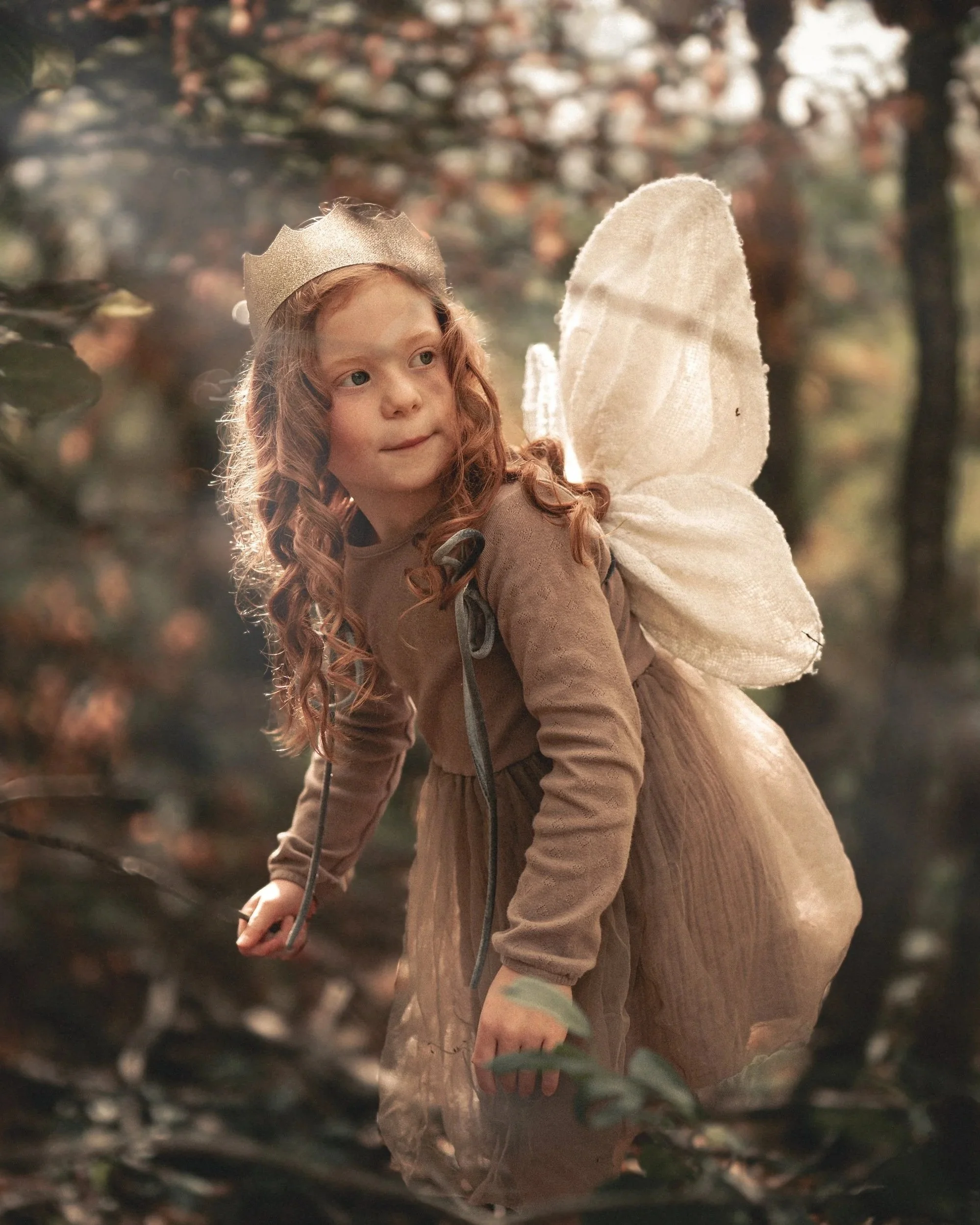 A young red headed girl with fairy wings and a gold crown moves through the forest, a ray of light makes her look like she's glowing