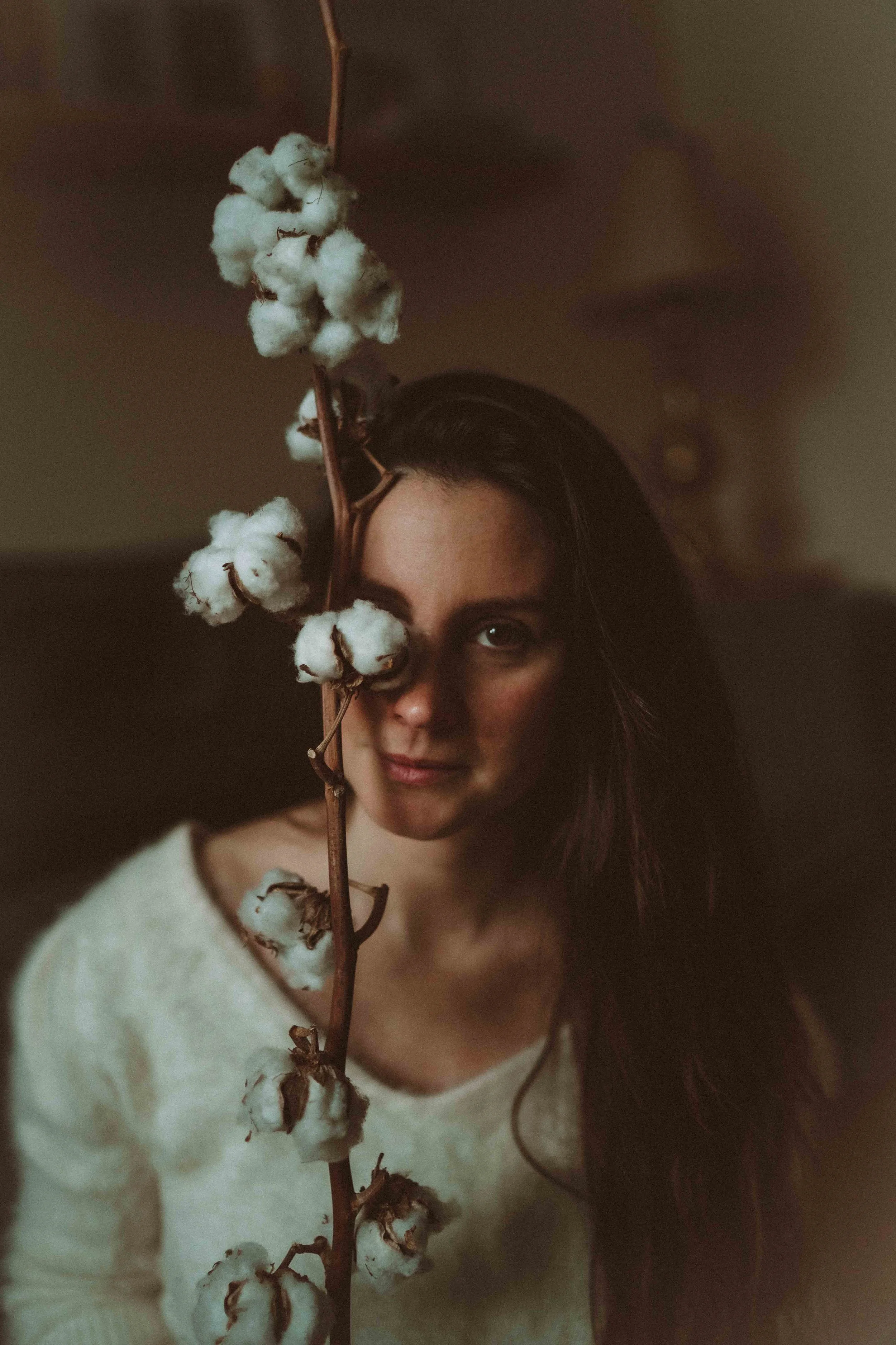 A woman with long dark hair sitting indoors, holding a cotton branch obstructing part of her face, wearing a light-colored sweater, with a neutral expression.