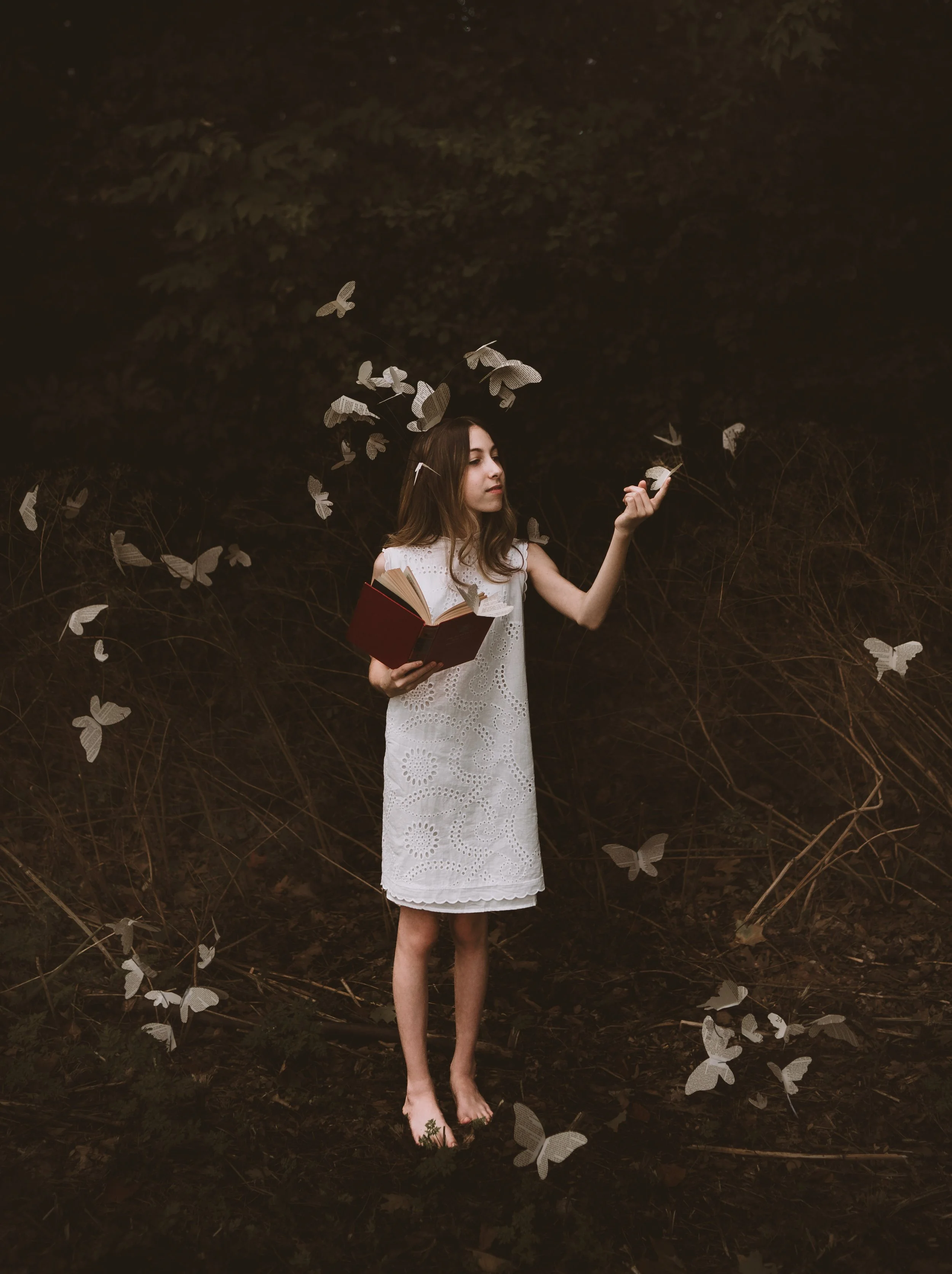 A young woman with brown hair in a white eyelet dress stands barefoot outdoors in a dark forest setting. She holds an open book in one hand and appears to be releasing paper butterflies from her other hand, with paper butterflies flying around her.