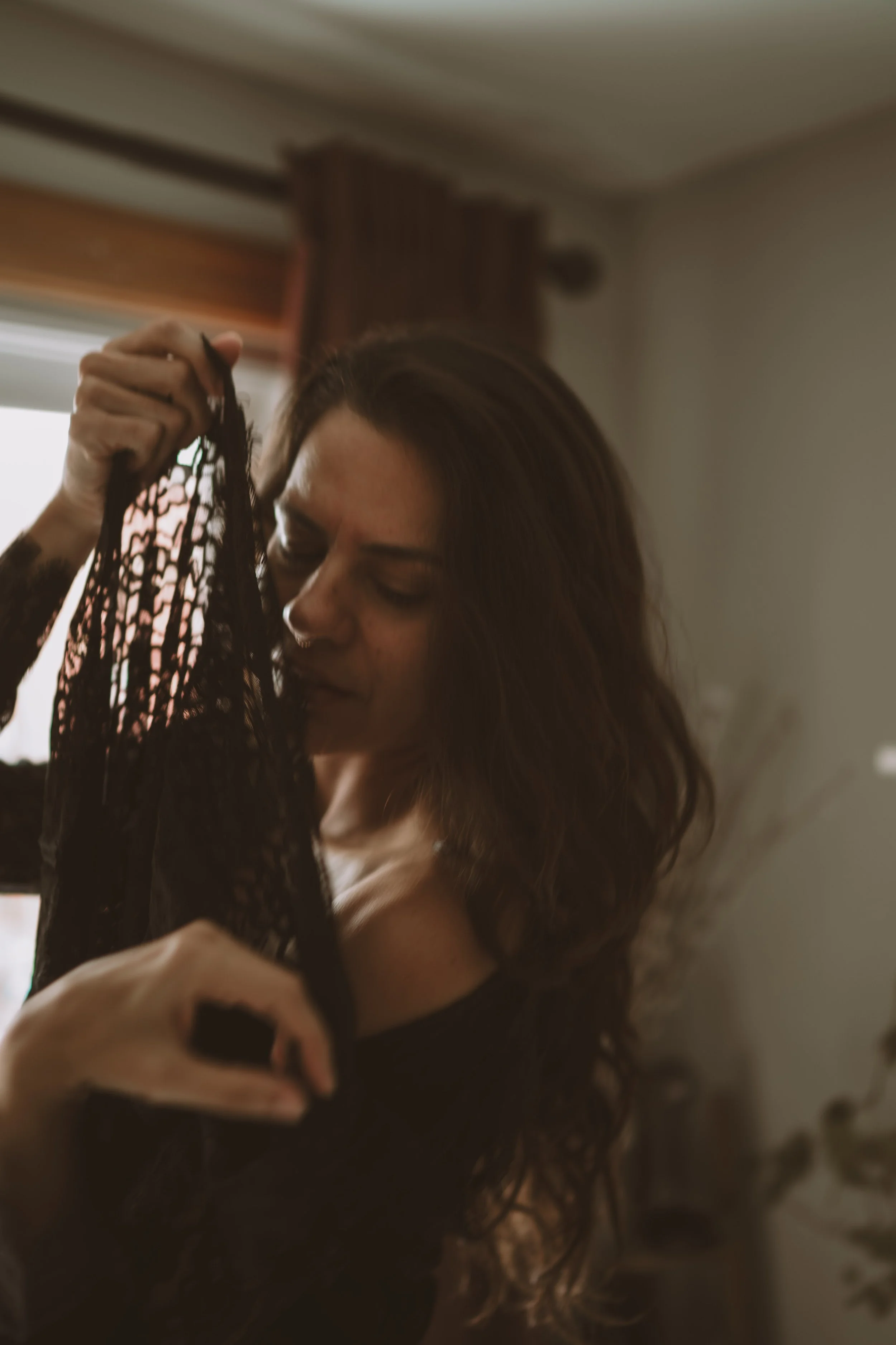 A woman with long wavy dark hair holding black lace lingerie close to her face indoors.