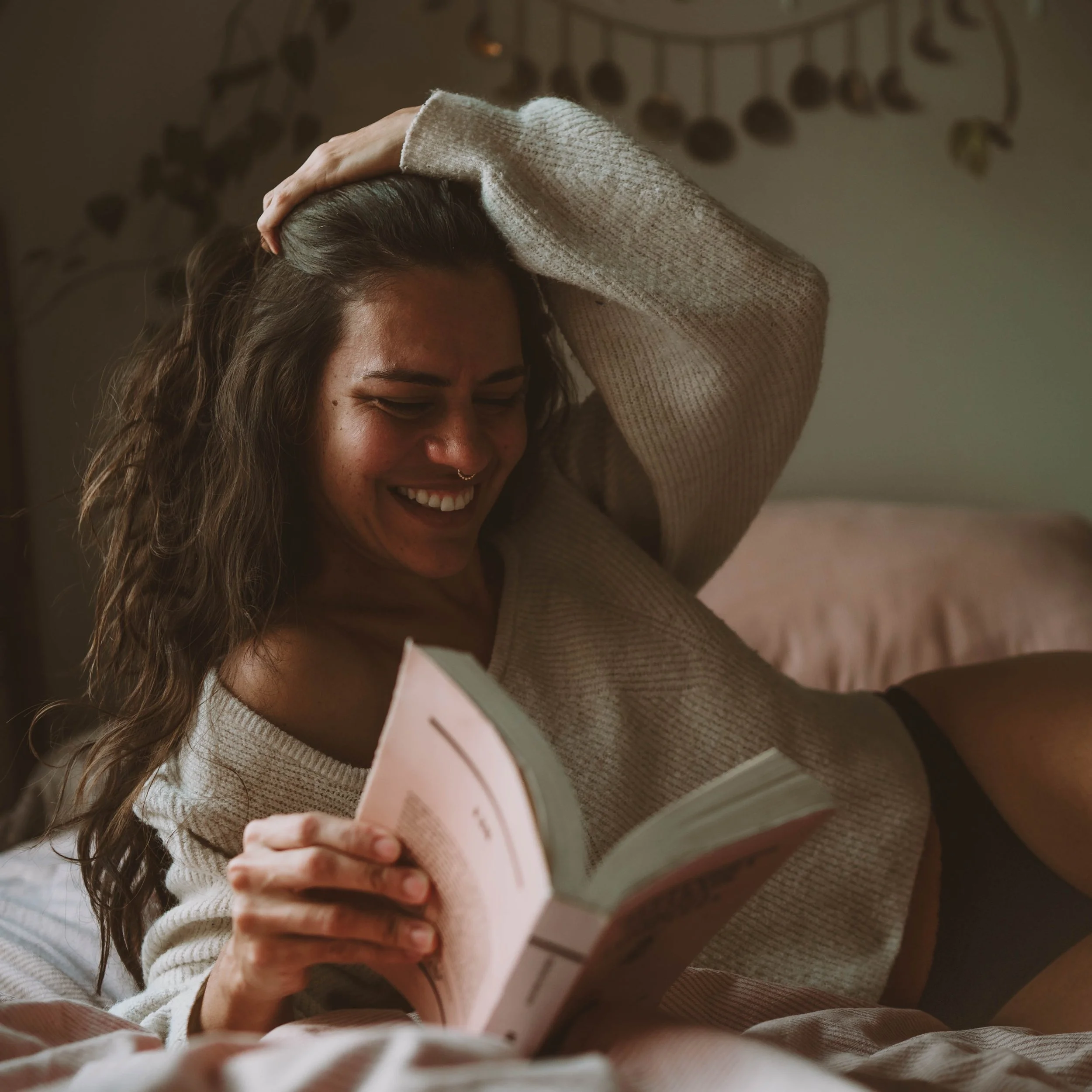 A woman with long curly hair and a septum piercing, wearing a beige sweater, is smiling while reading a book, sitting on a bed in a cozy room.