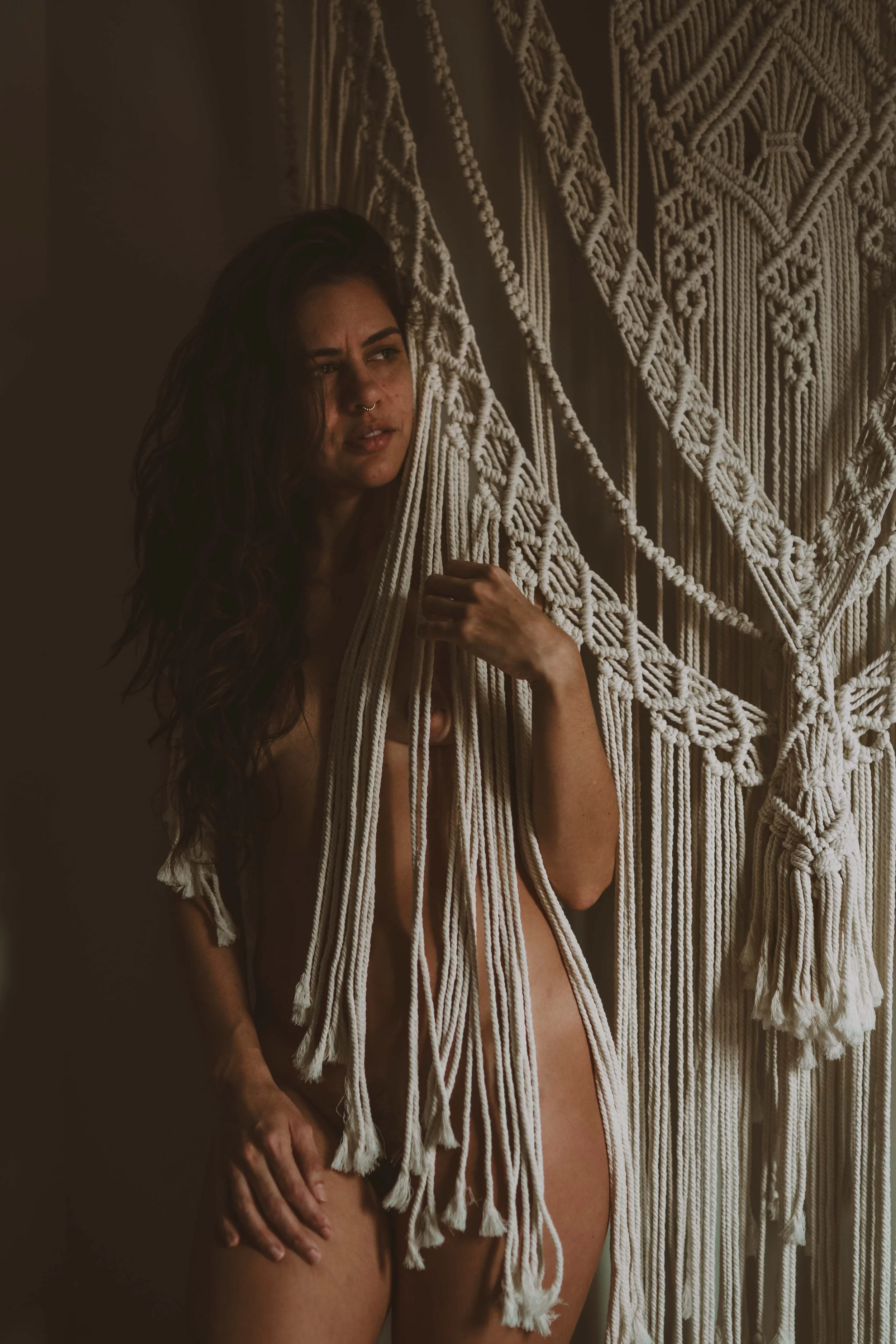 A woman with long dark hair and a septum piercing standing partially nude next to a large, intricately woven macramé wall hanging.