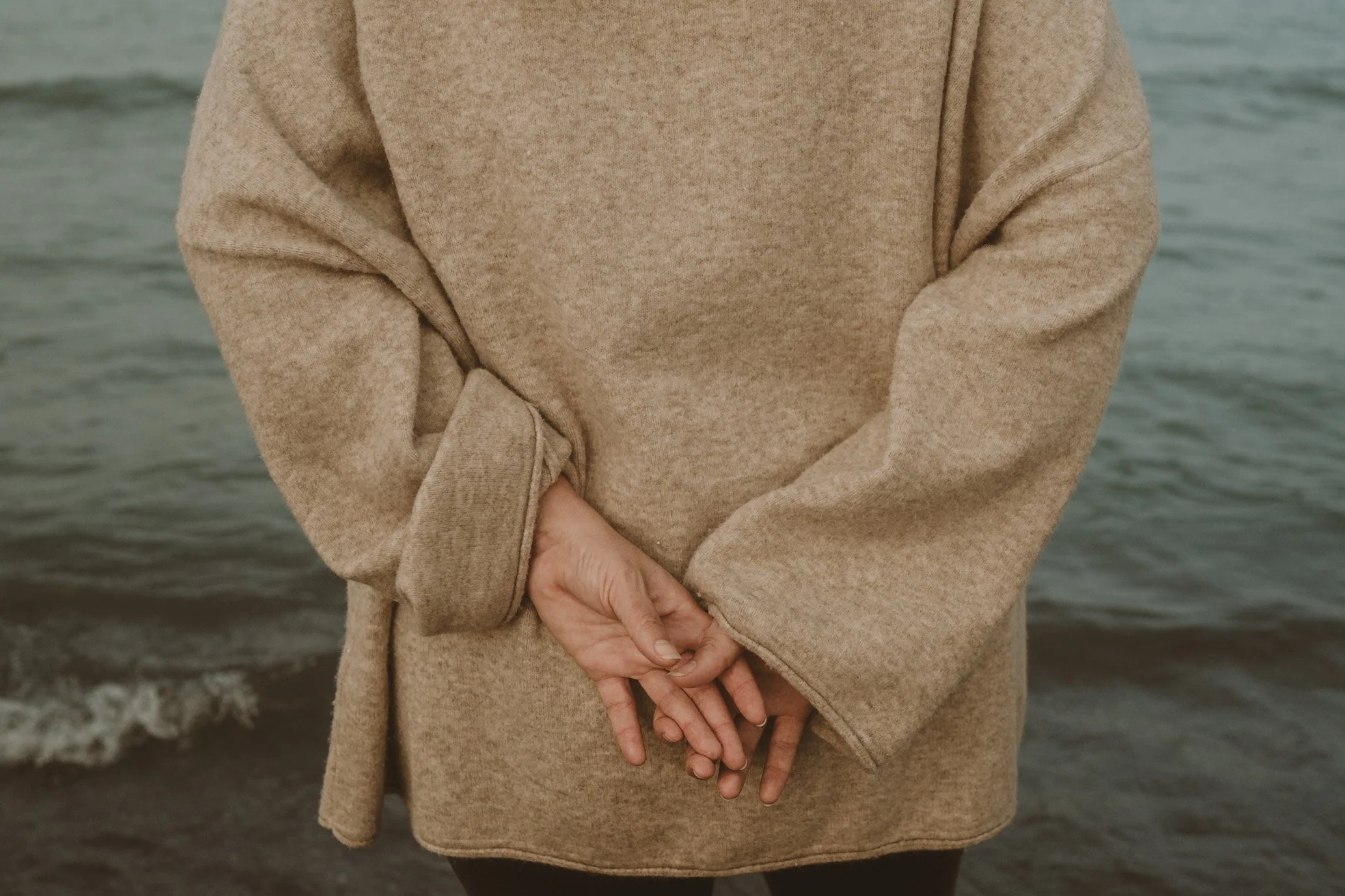 Person wearing a beige sweater with their hands folded in front of them, standing near a body of water.