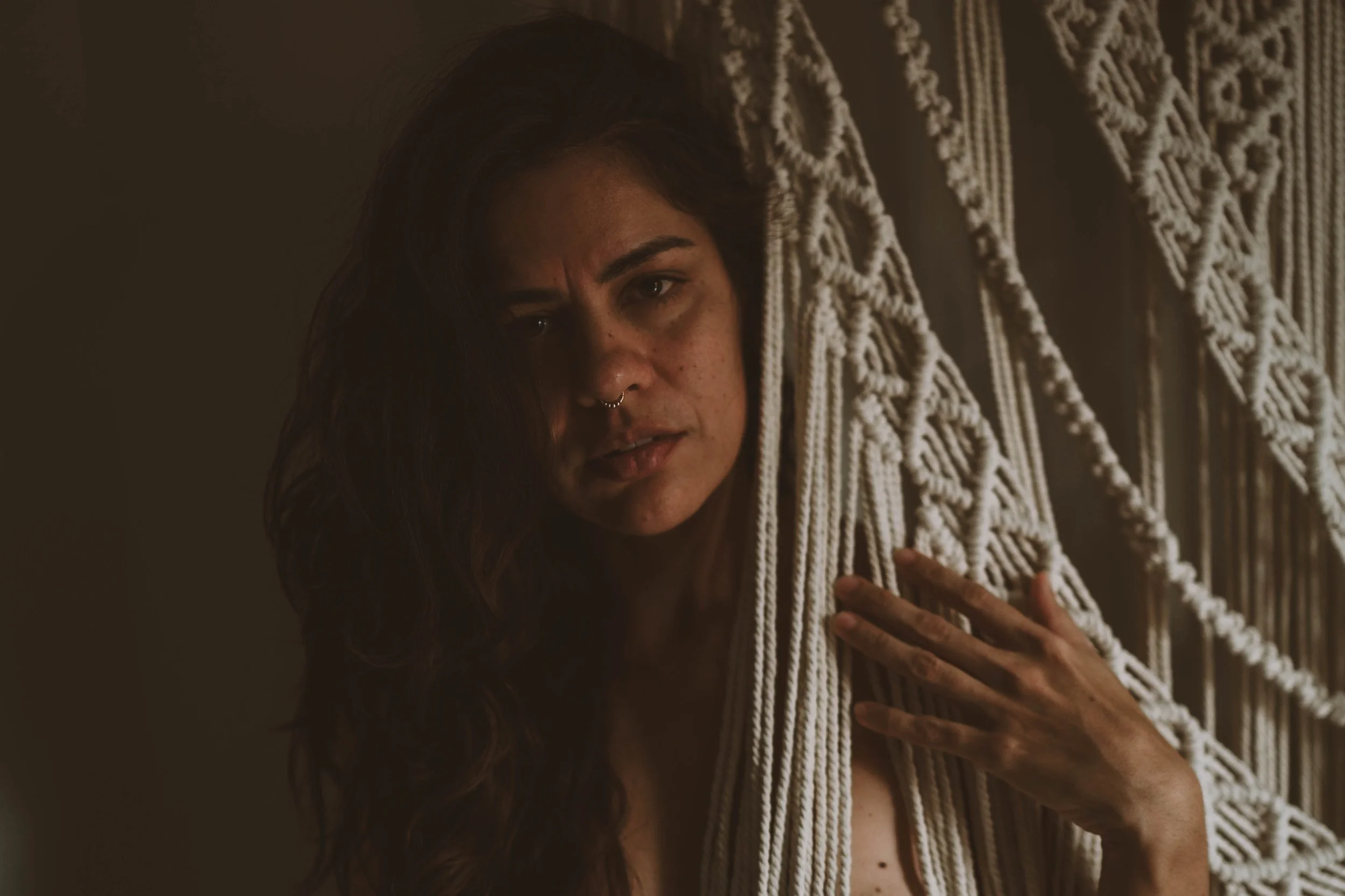A woman with long dark hair, wearing a septum piercing, looks distressed while holding a macrame wall hanging or curtain.