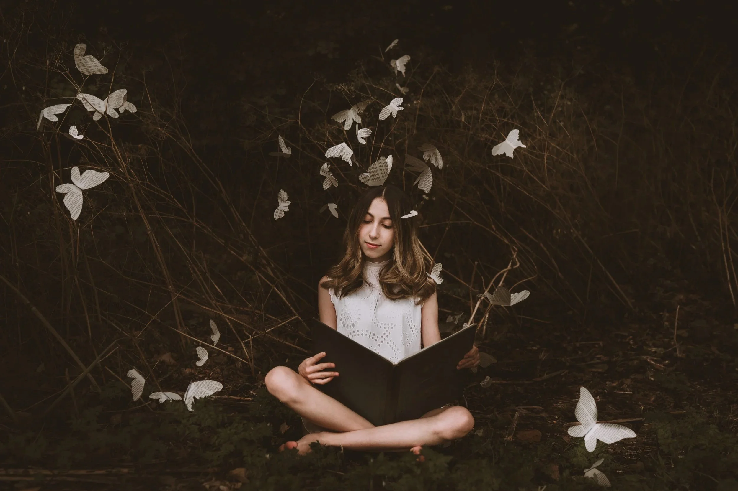 A young woman with long wavy hair sitting cross-legged outdoors on the ground, immersed in reading a book. She is surrounded by a dark, natural setting with leafless branches and numerous butterfly-shaped paper cutouts hanging around her, some floating in the air.