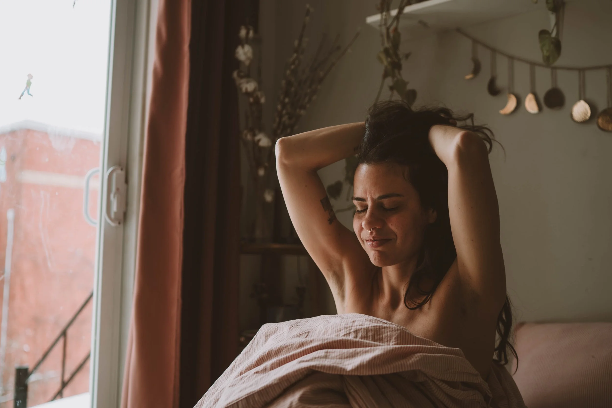 A woman with dark curly hair stretching with her arms raised above her head in bed, near a window with pink curtains, bright daylight, and decorative wall hanging behind her.