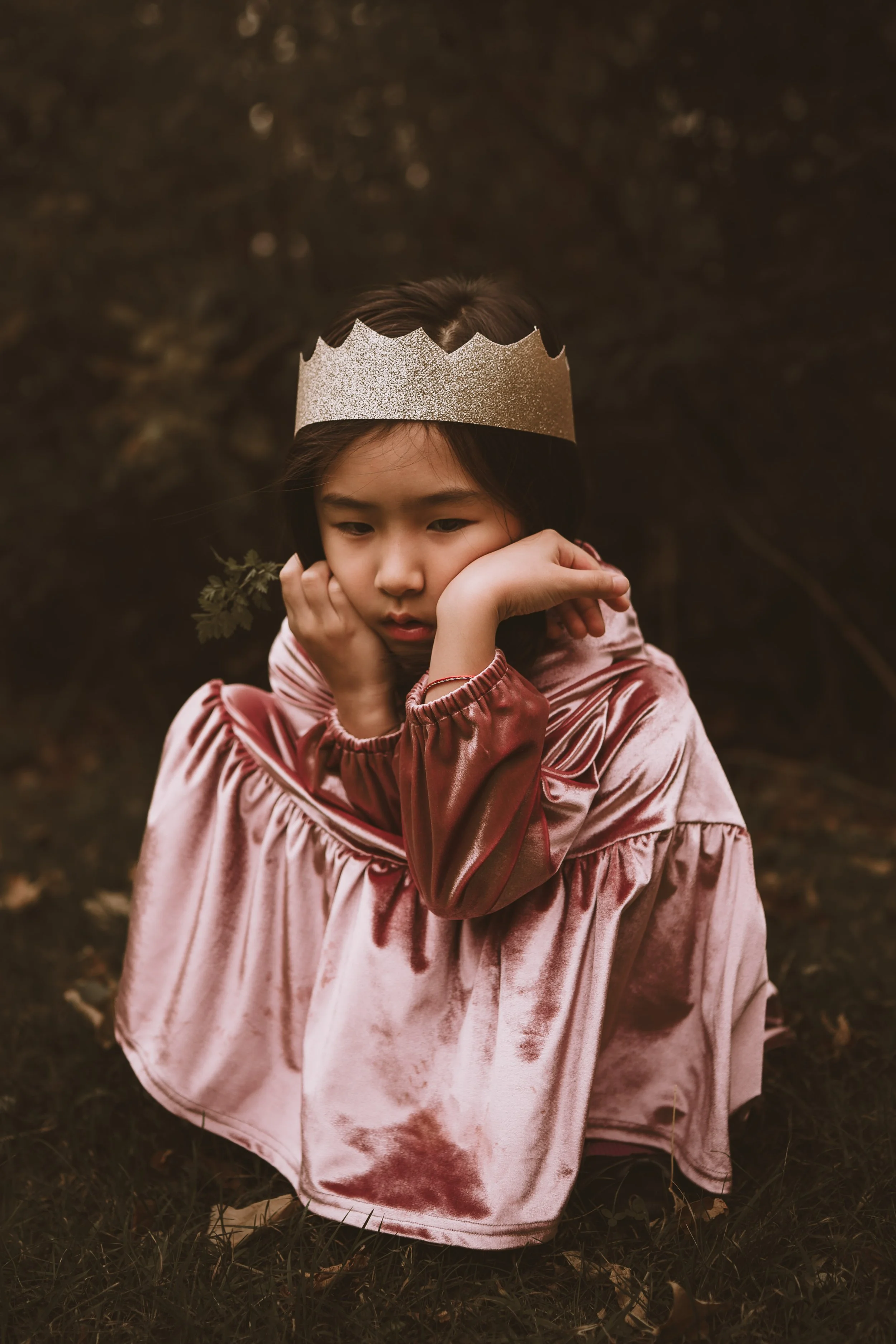 A young girl sitting outdoors on the ground with brown leaves and grass, wearing a large pink tie-dye hoodie and a glittery silver crown, resting her chin on her hands and looking downward.