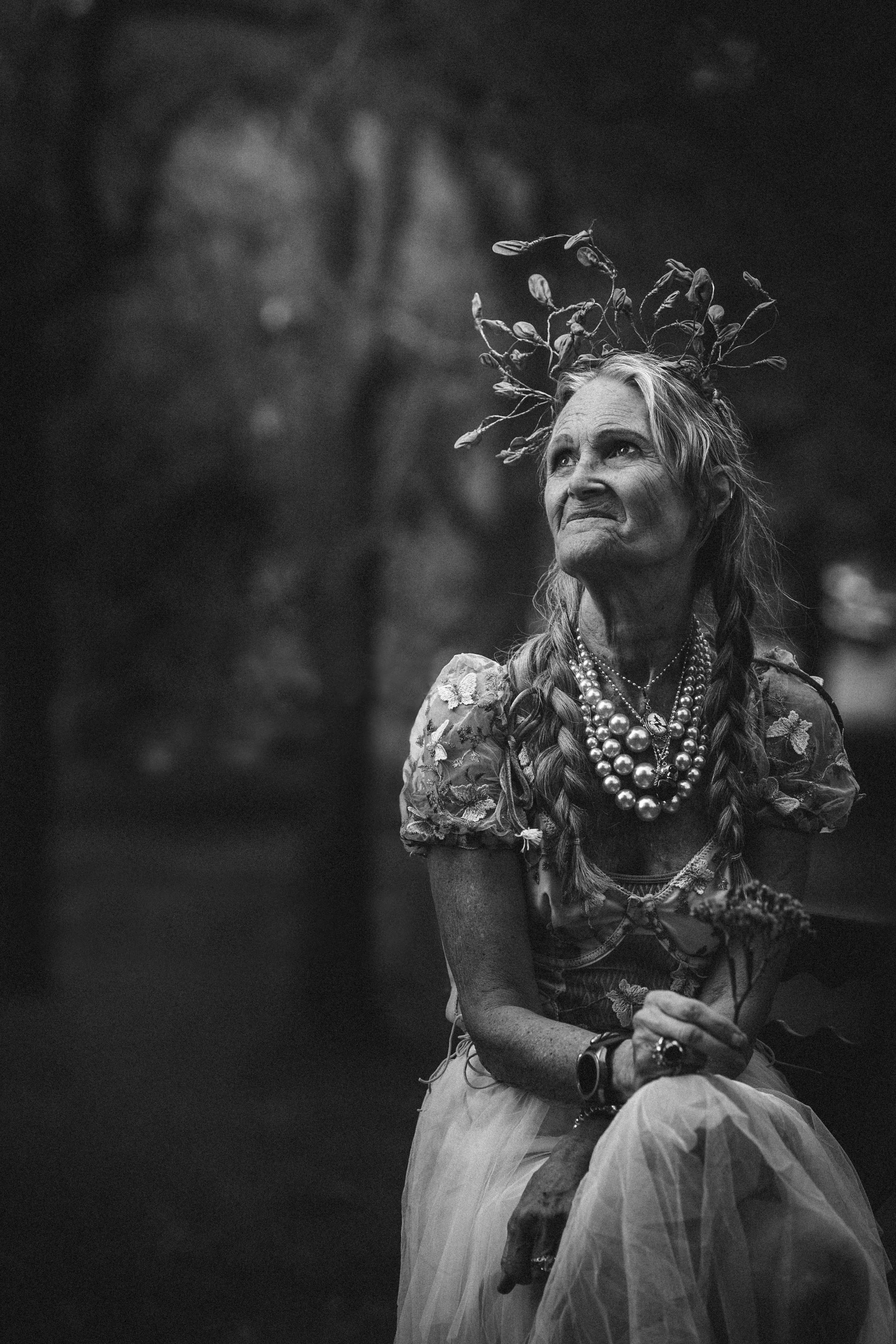 Black and white photo of an elderly woman with long braids, wearing multiple pearl necklaces and a floral embroidered dress, sitting outdoors with a serene and contemplative expression.