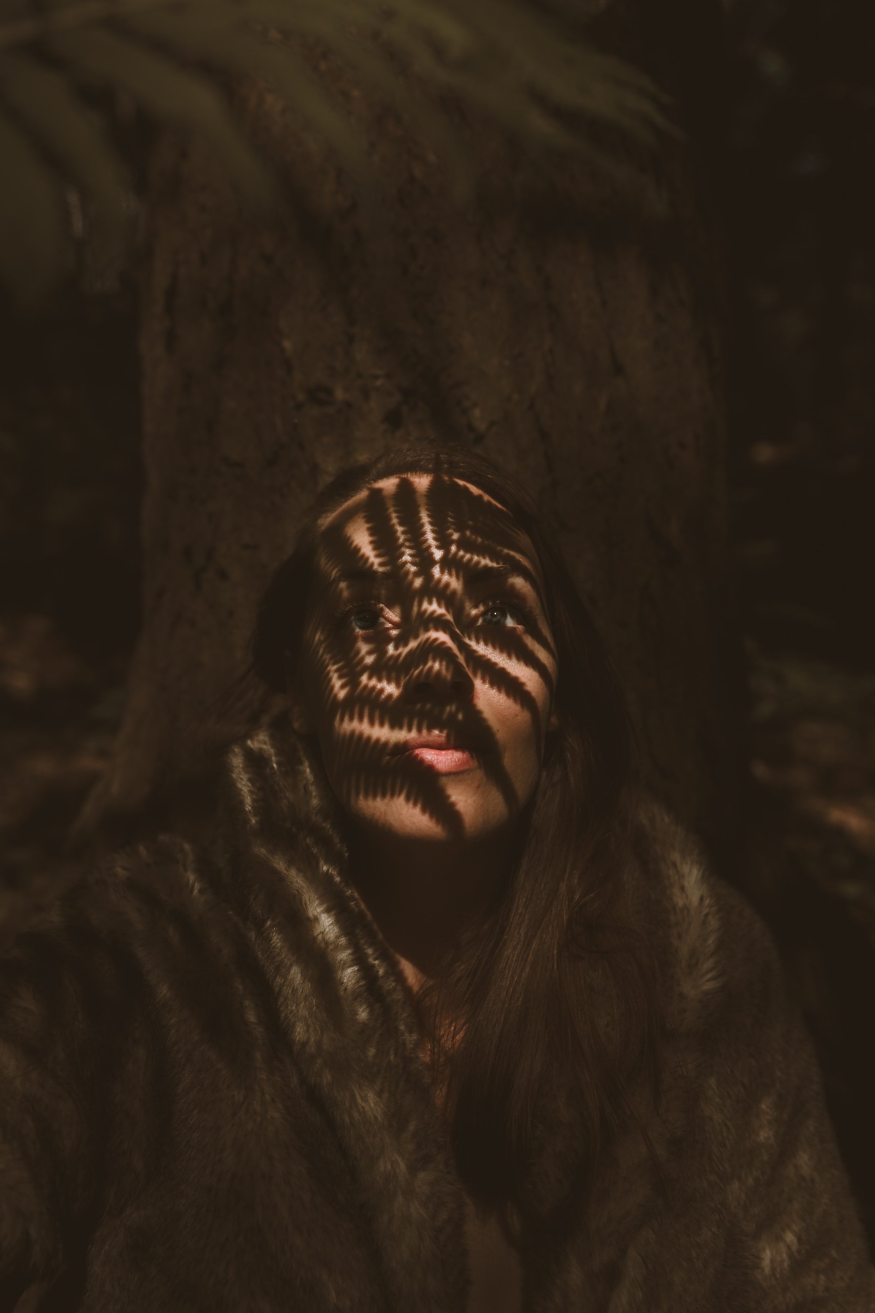 A young woman with long hair, partially covered in a fur coat, looking up into the camera with shadows cast across her face by nearby objects, creating a striped pattern.
