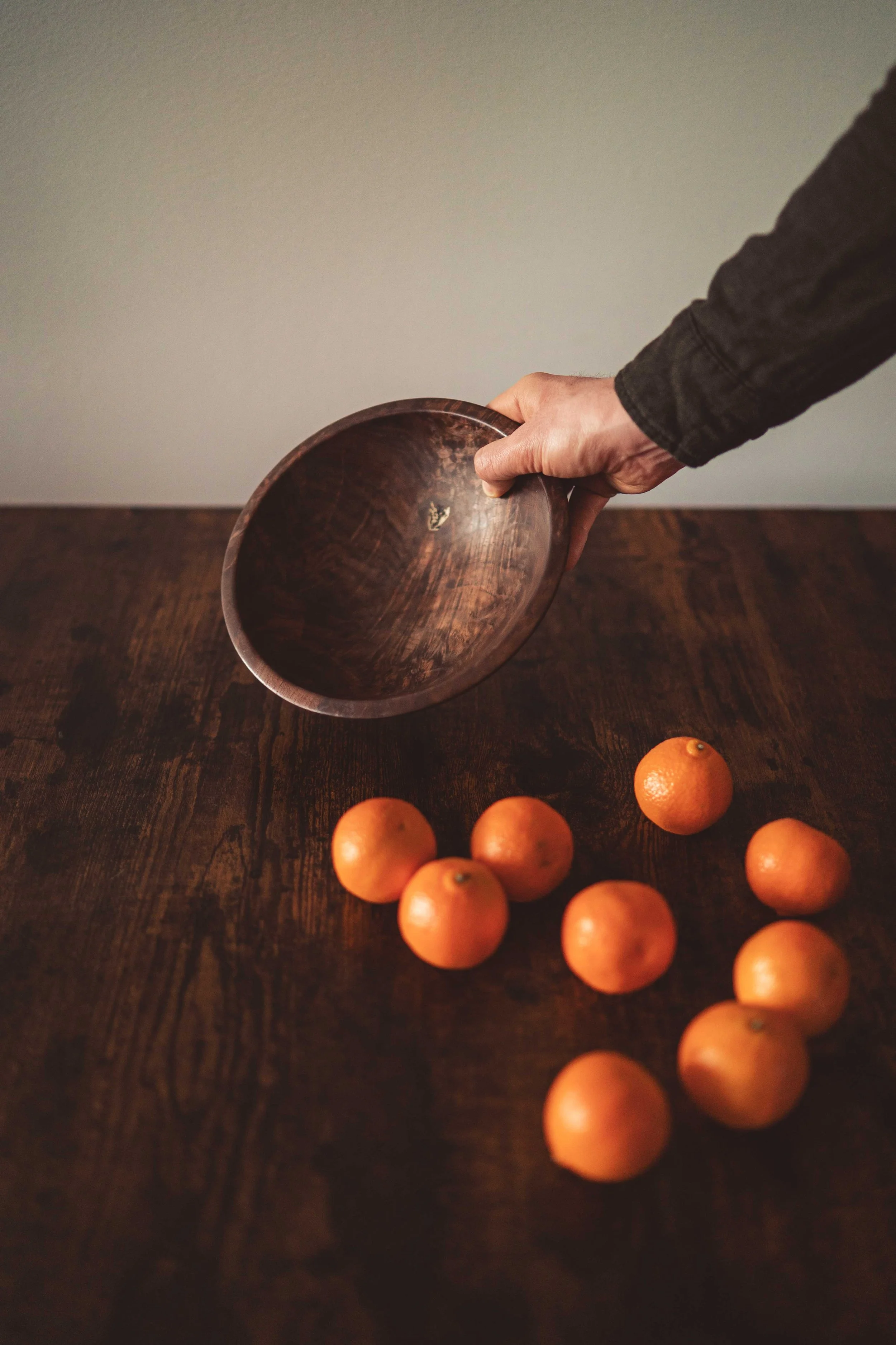 A hand holding a wooden bowl over a dark wooden table with nine oranges scattered on it.