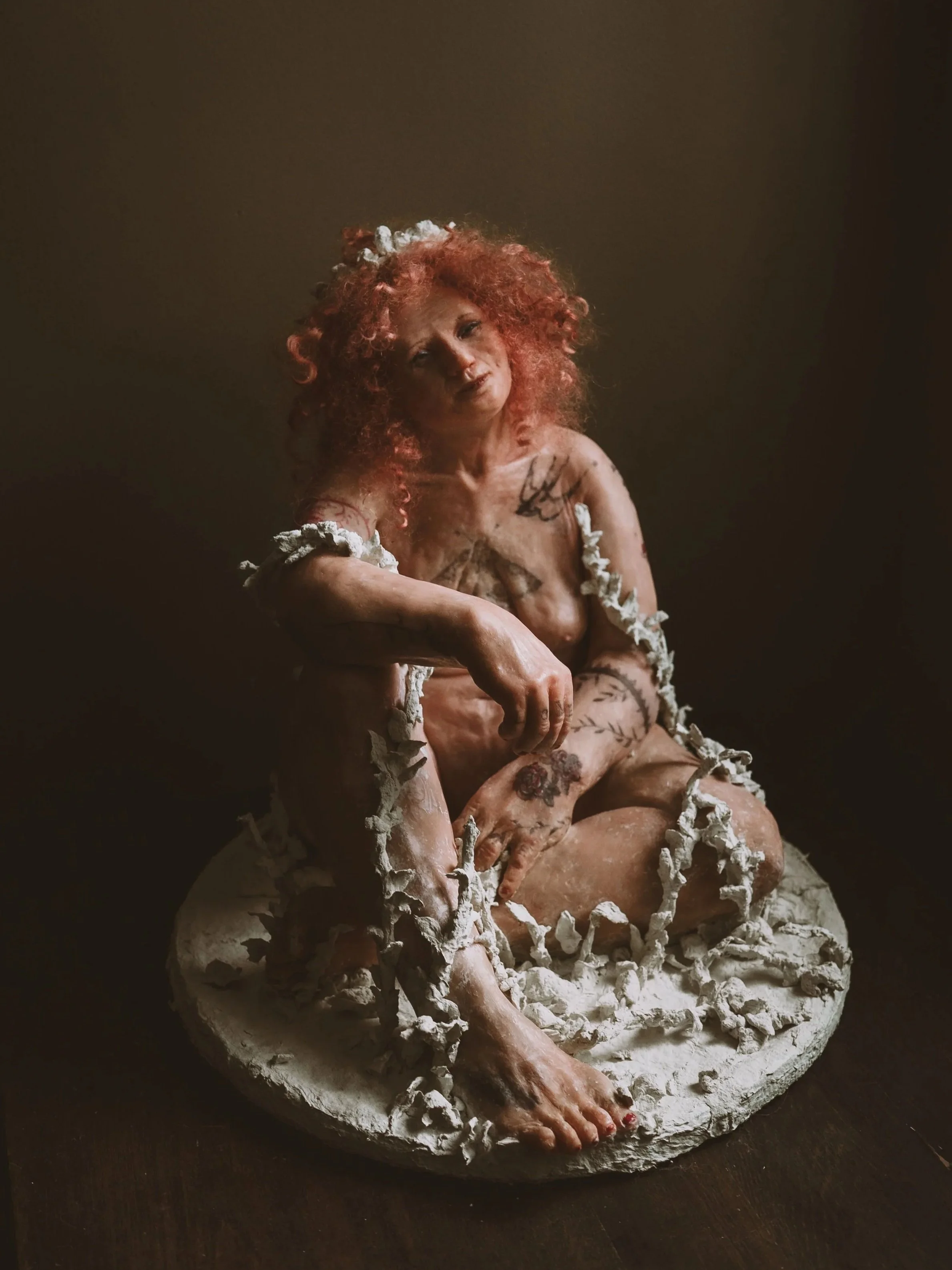 A woman with curly red hair sitting on a textured white platform, surrounded by white decorative elements, against a dark background.