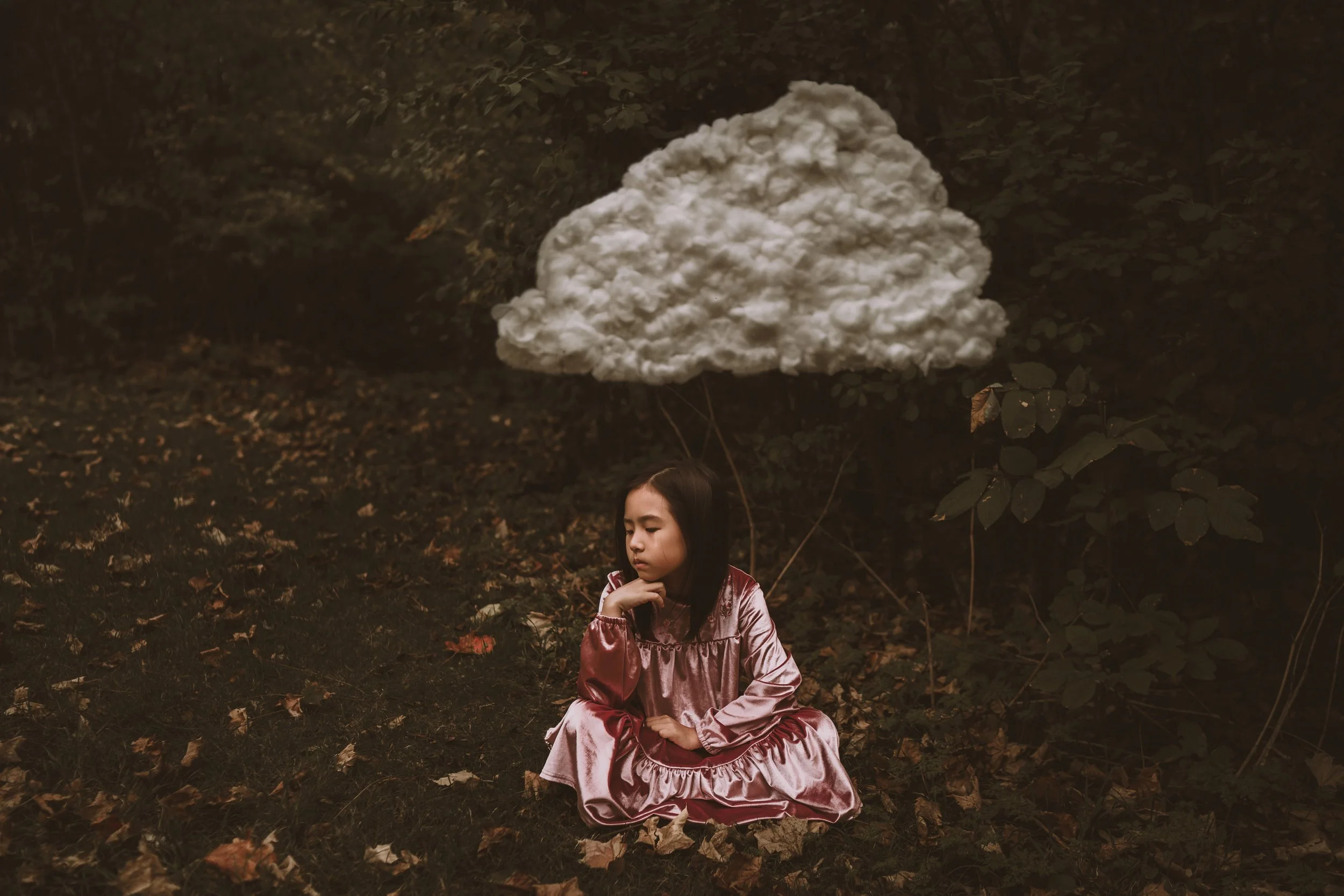 A young girl sitting on the ground in a forest, wearing a pink satin dress, with her eyes closed and hand near her neck, beneath a floating, glowing cloud-shaped object in a dark, leafy environment.