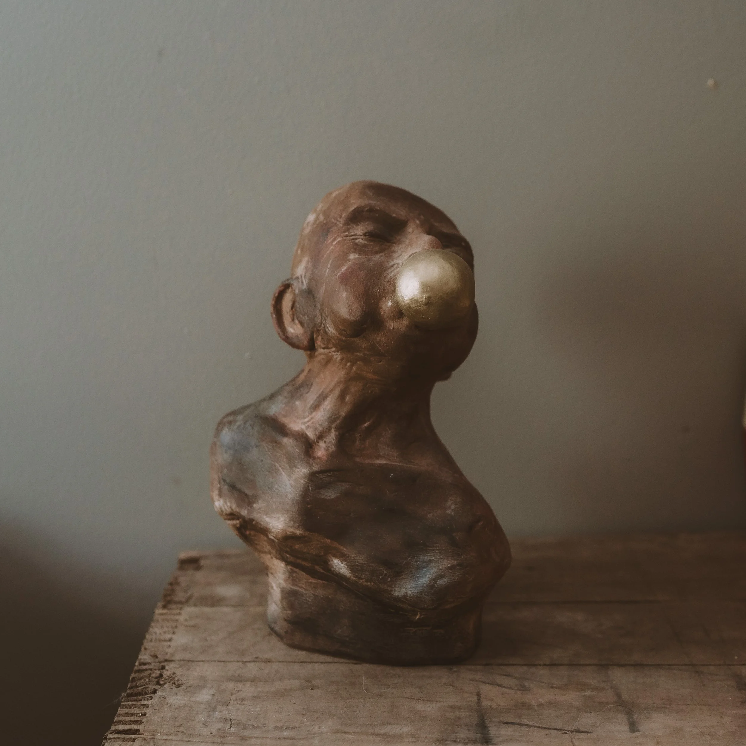 Close-up of a carved wooden sculpture of a smiling face with a rounded nose, placed on a wooden surface with a textured gray background.