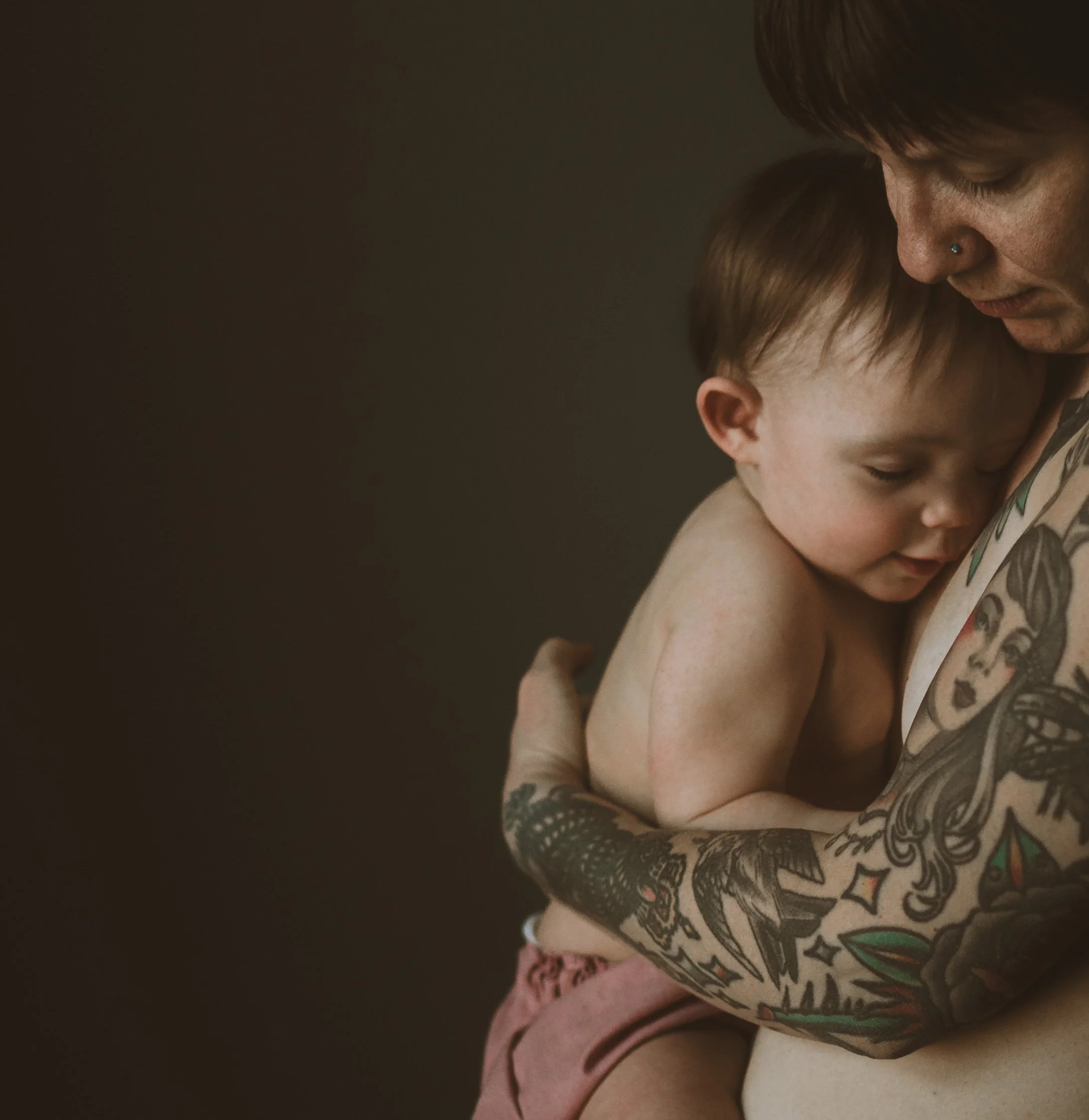Close-up of a mother and young girl, both with eyes closed, leaning their foreheads together, showing a tender moment of connection, with the mom wearing a nose ring and the girl having tattoos on her arm.