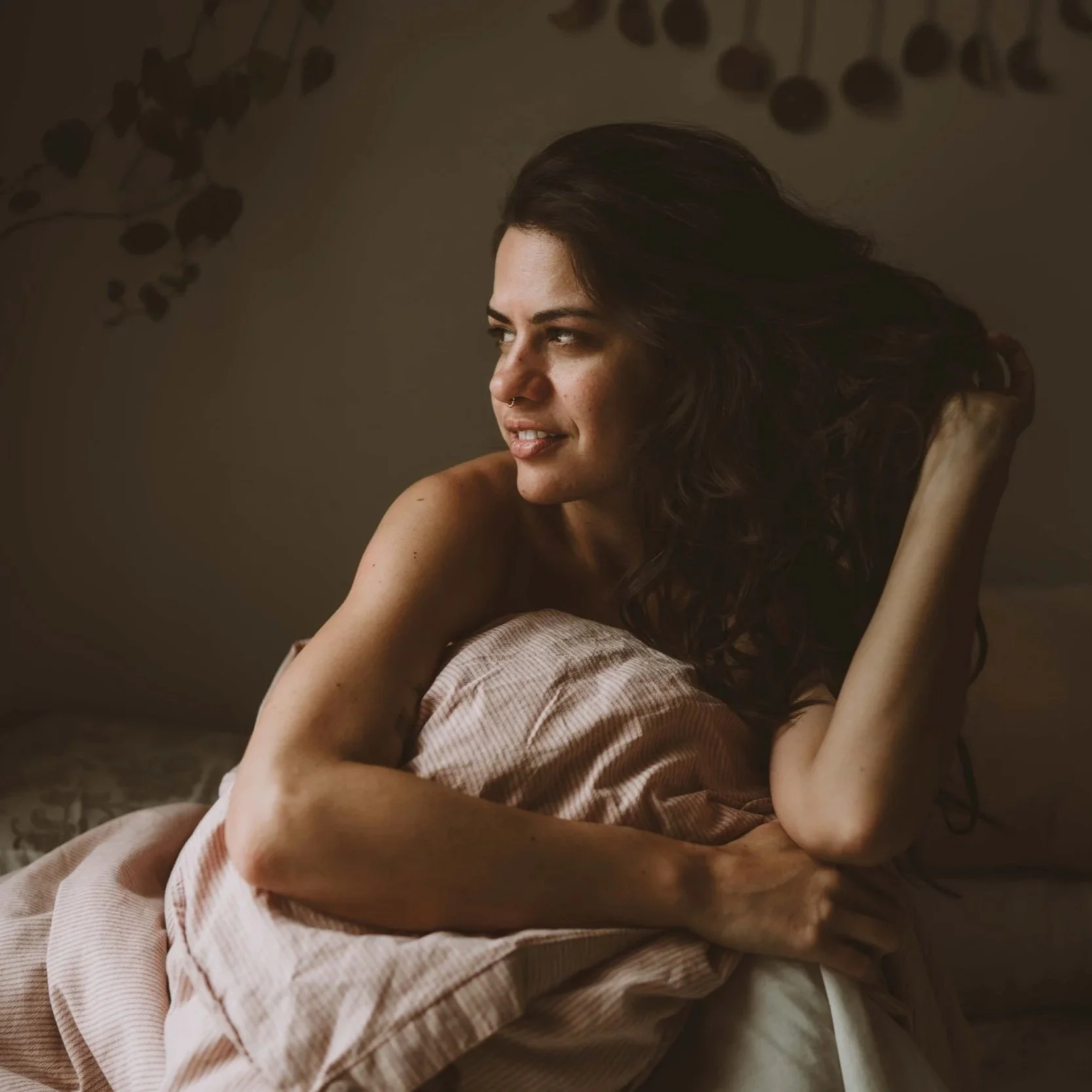 A woman with dark hair and a nose ring laying on a bed, gazing to her left with a thoughtful expression, in soft natural lighting.