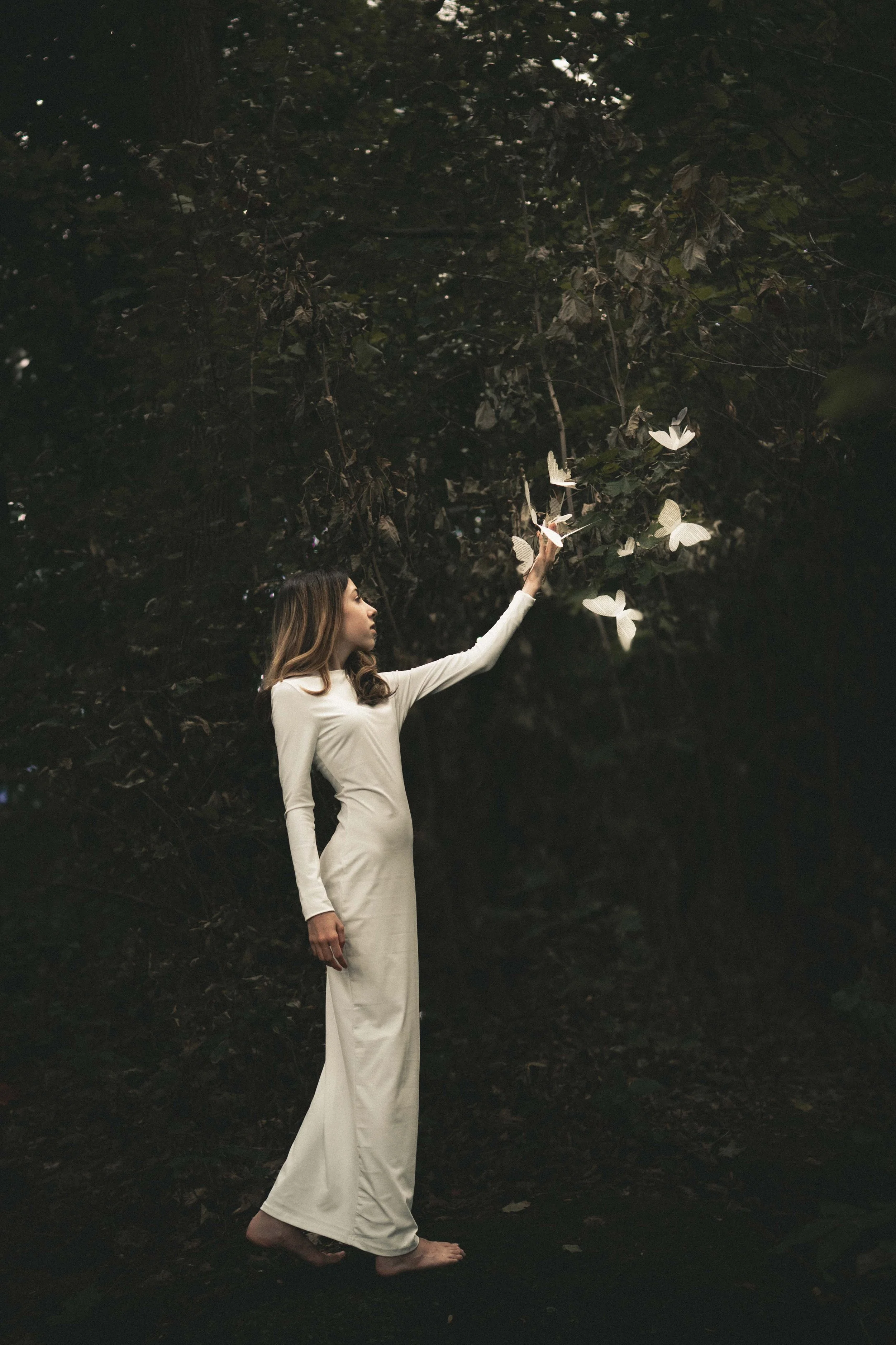 A woman in a long white gown touching floating glowing butterflies in a dark forest.