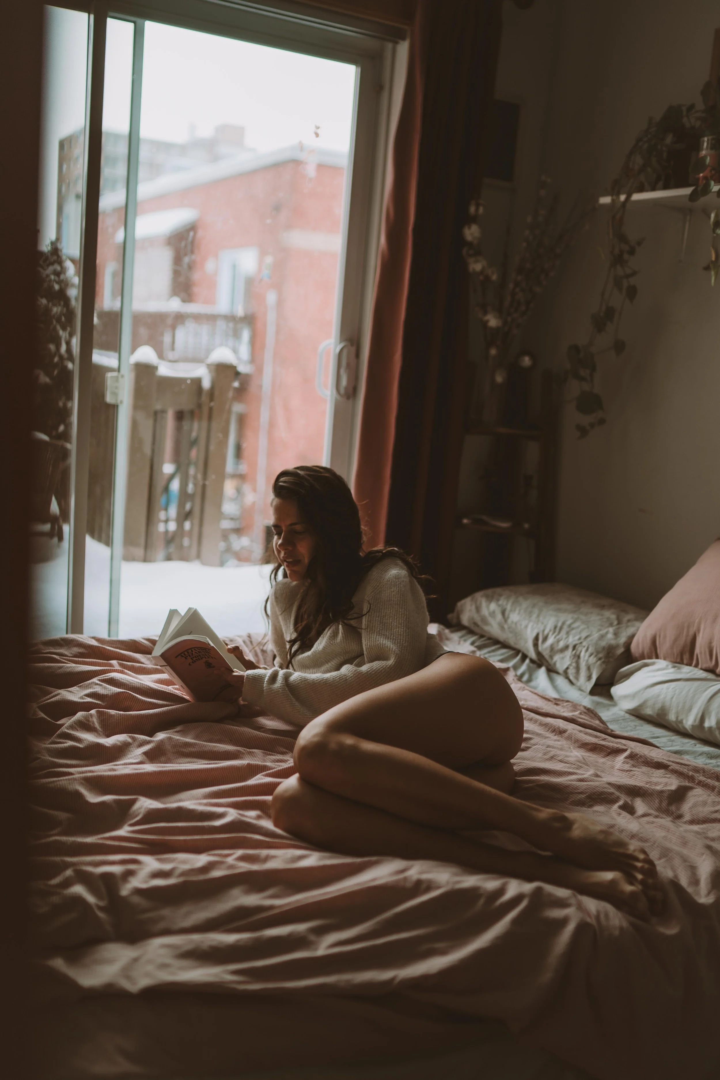 A woman with long dark hair lies on her stomach on a bed, reading a book, with a window showing a snowy outdoor scene in the background.