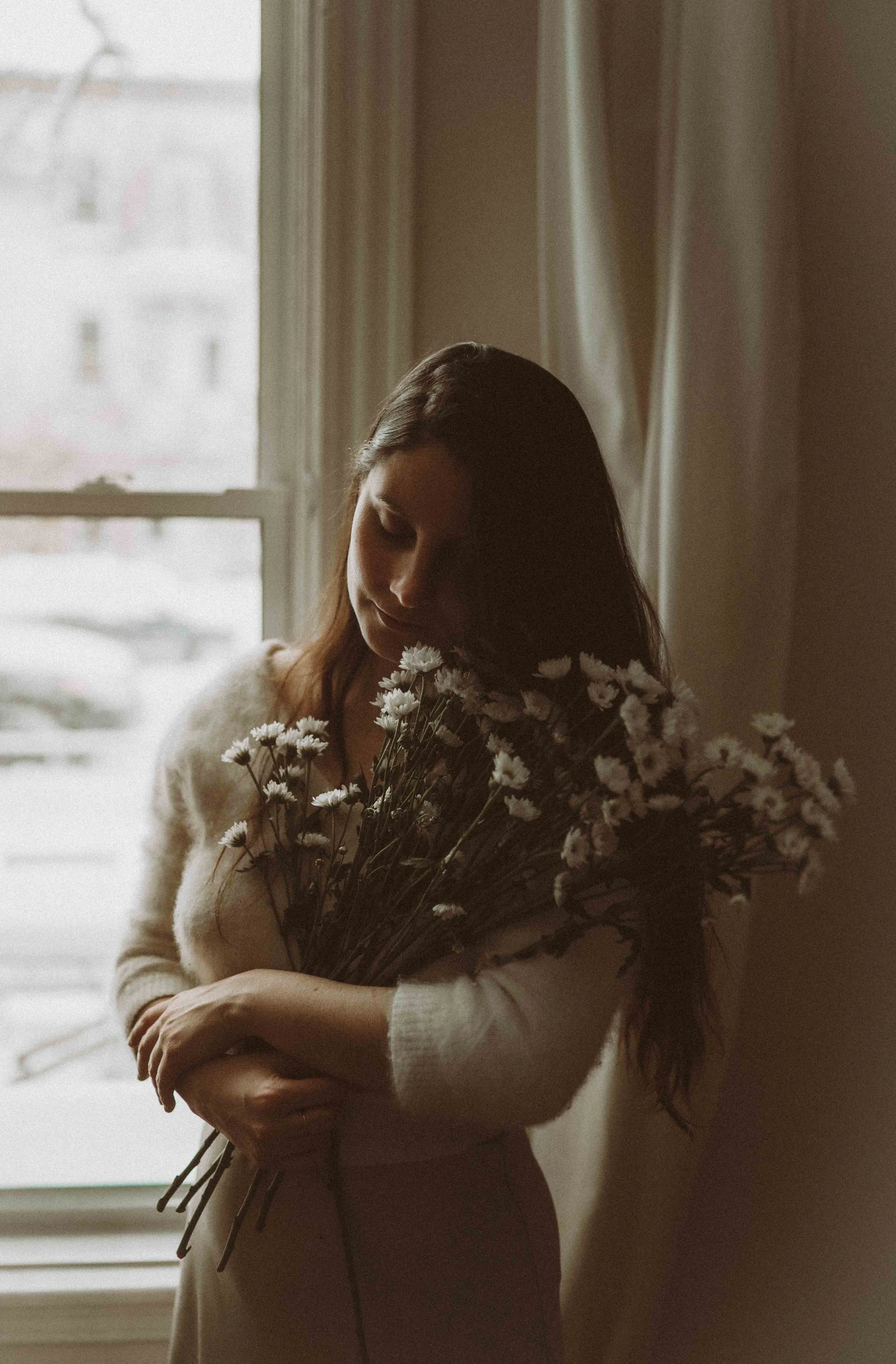 A woman holding a bouquet of white flowers near a window with curtains, soft natural light illuminating her face.