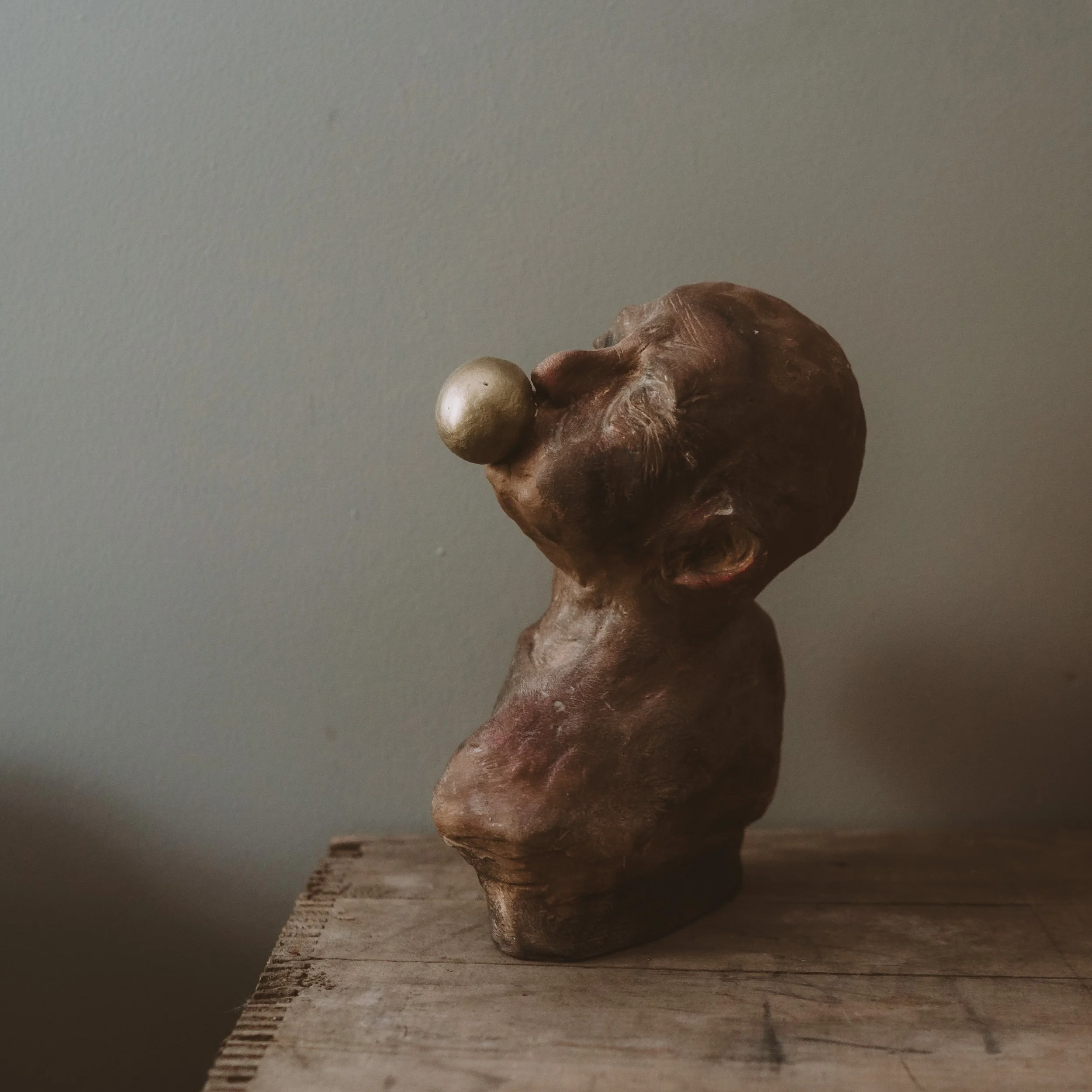 Close-up of a small, textured, brown, irregularly shaped sculpture with a silver sphere on top, placed on a wooden surface against a plain background.