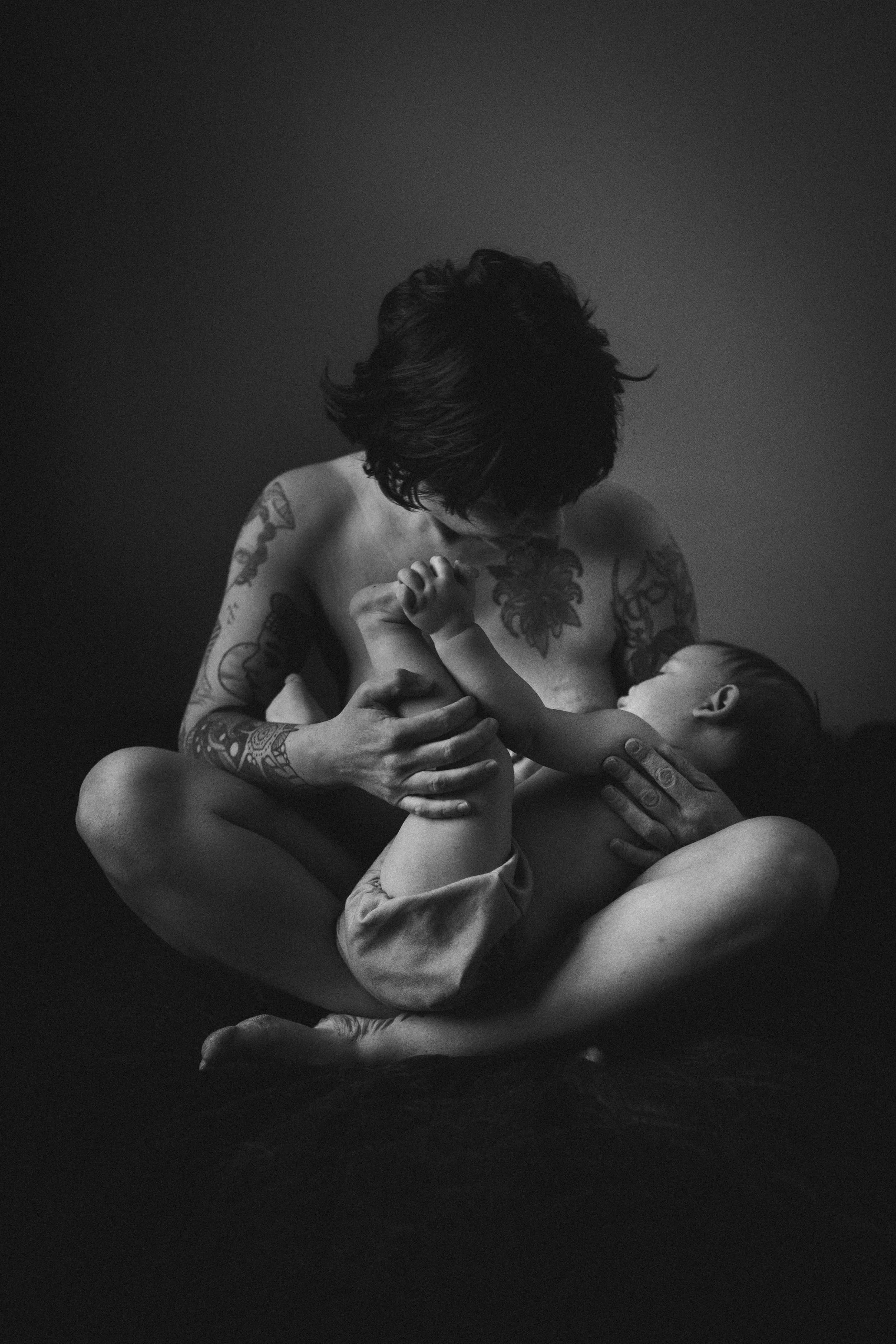 A black and white photo of a tattooed woman with short hair holding a baby close to her chest, both sitting with their legs crossed, against a dark background.