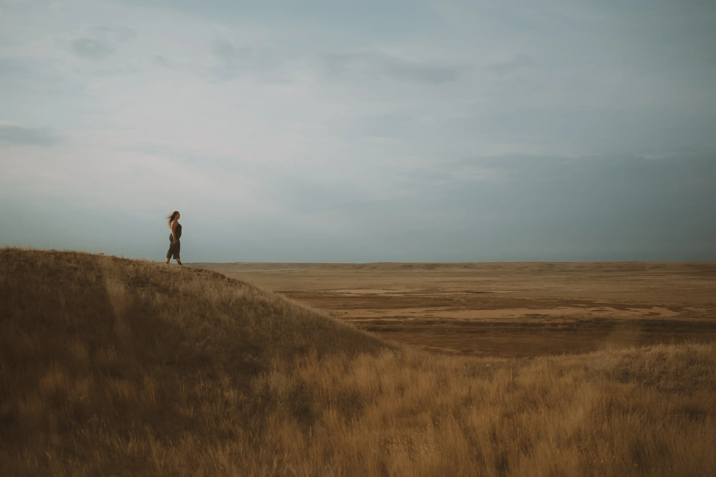 A woman with long hair standing alone on a grassy cliff overlooking a barren plain under a cloudy sky.