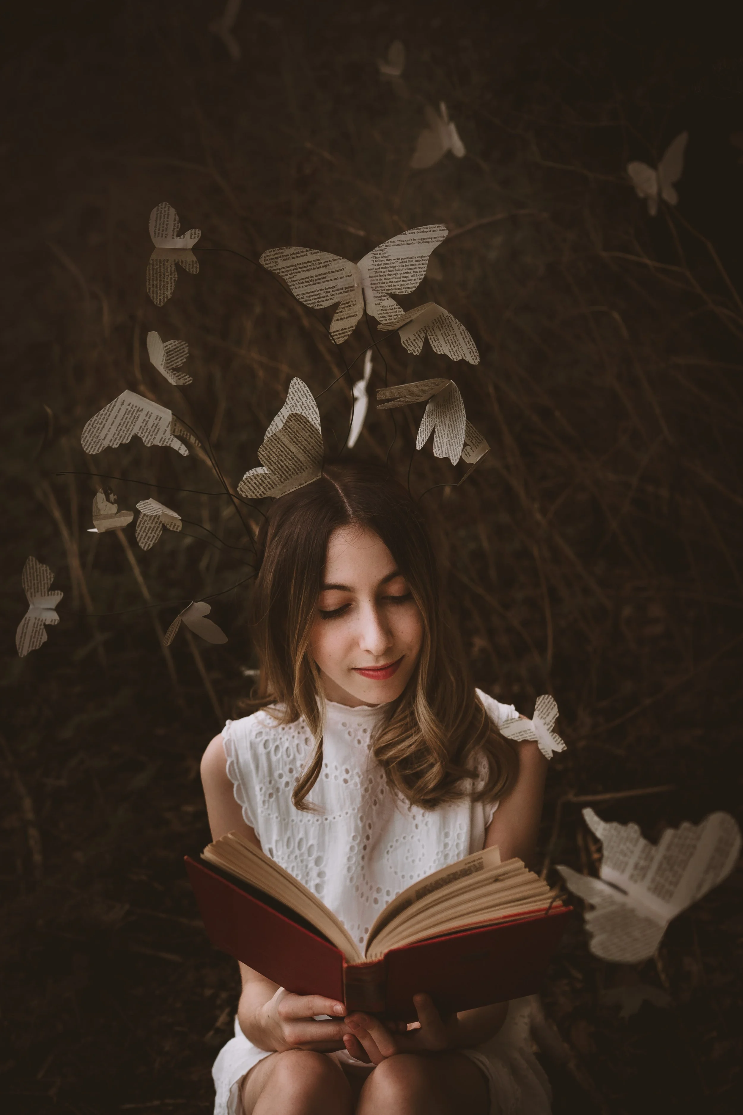 A young woman with light brown, wavy hair, wearing a white eyelet dress, sitting outdoors against a dark background with trees. She is holding an open red book and is surrounded by paper butterflies made from pages of text, some attached to thin wire