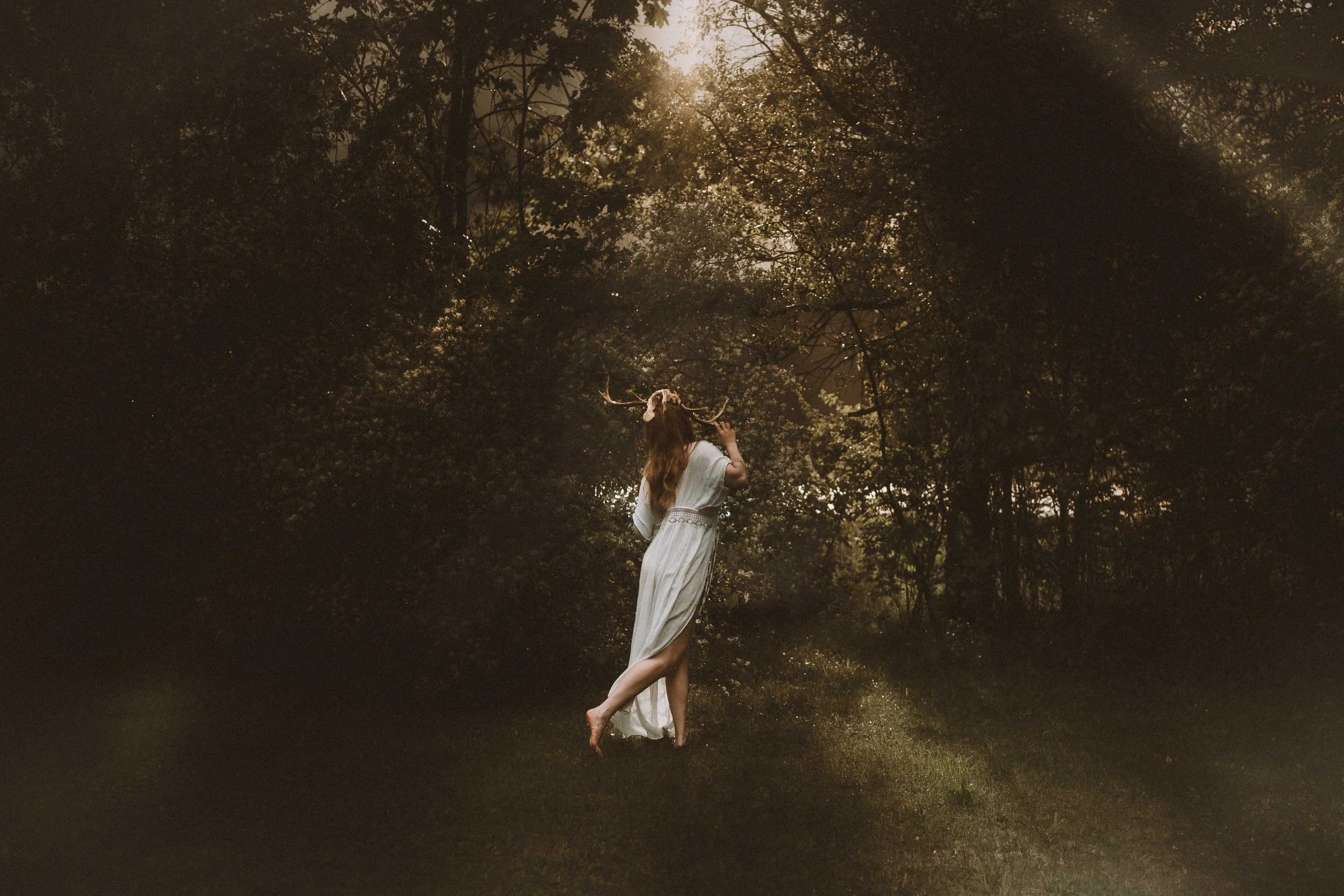 A person wearing a white flowing dress with deer antlers on their head stands in a forest clearing during sunset, with trees surrounding them and sunlight filtering through the leaves.