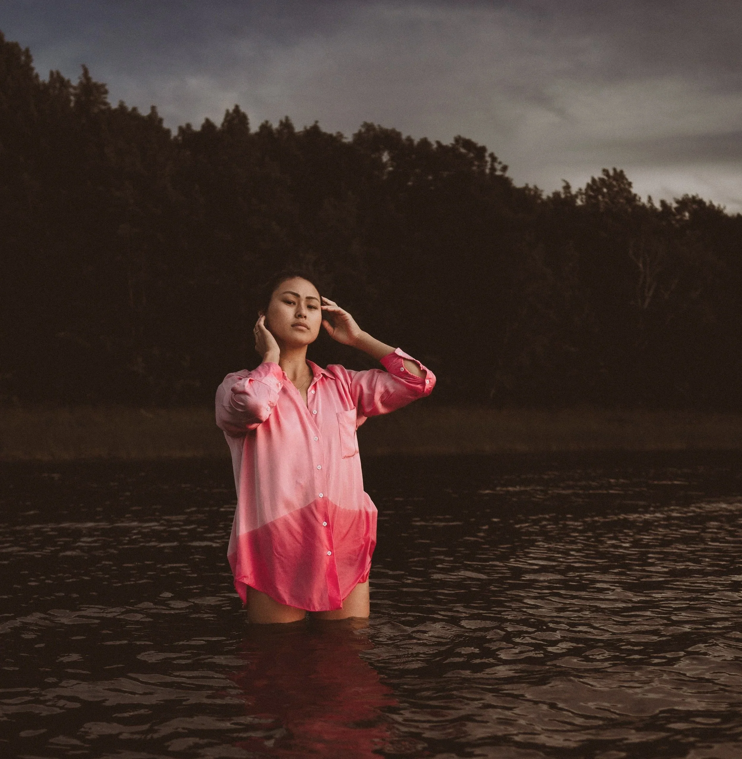 A woman in a pink shirt standing in a body of water with a dark, tree-covered hillside in the background, under a cloudy sky.