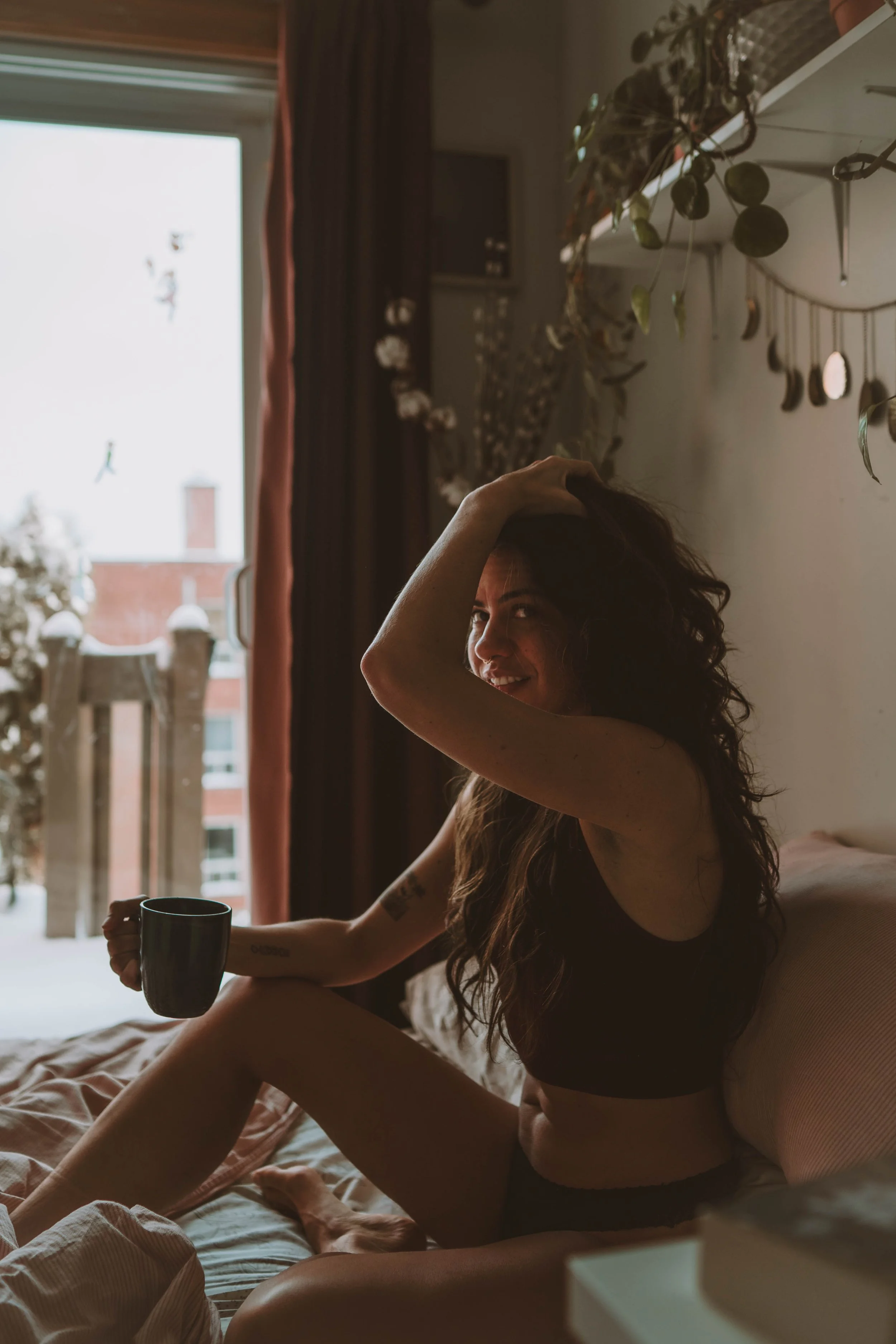 A woman sitting on a bed in a cozy room, holding a coffee mug, smiling, with a large window behind her showing a snowy outdoor scene.