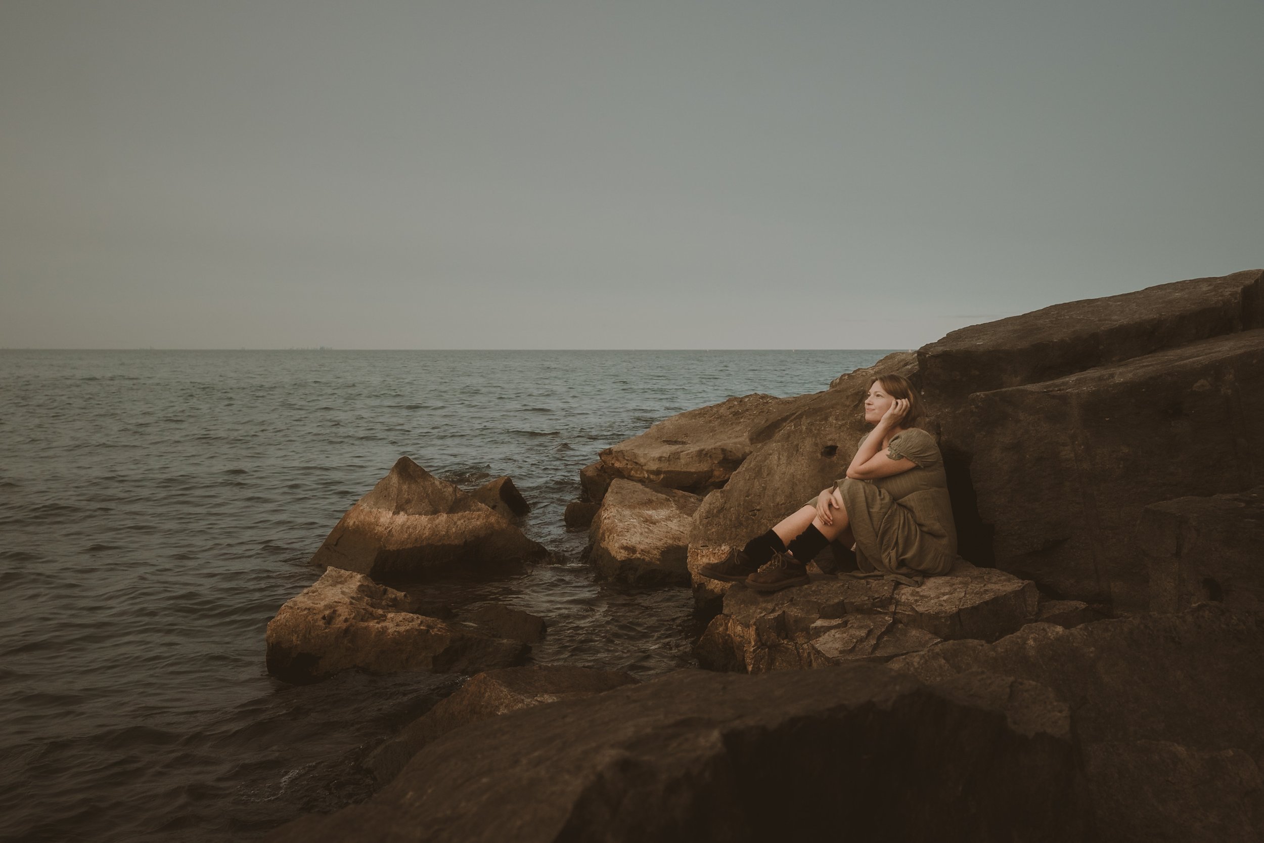 A woman sitting on rocks by the ocean, gazing thoughtfully at the water, with a calm and peaceful expression.